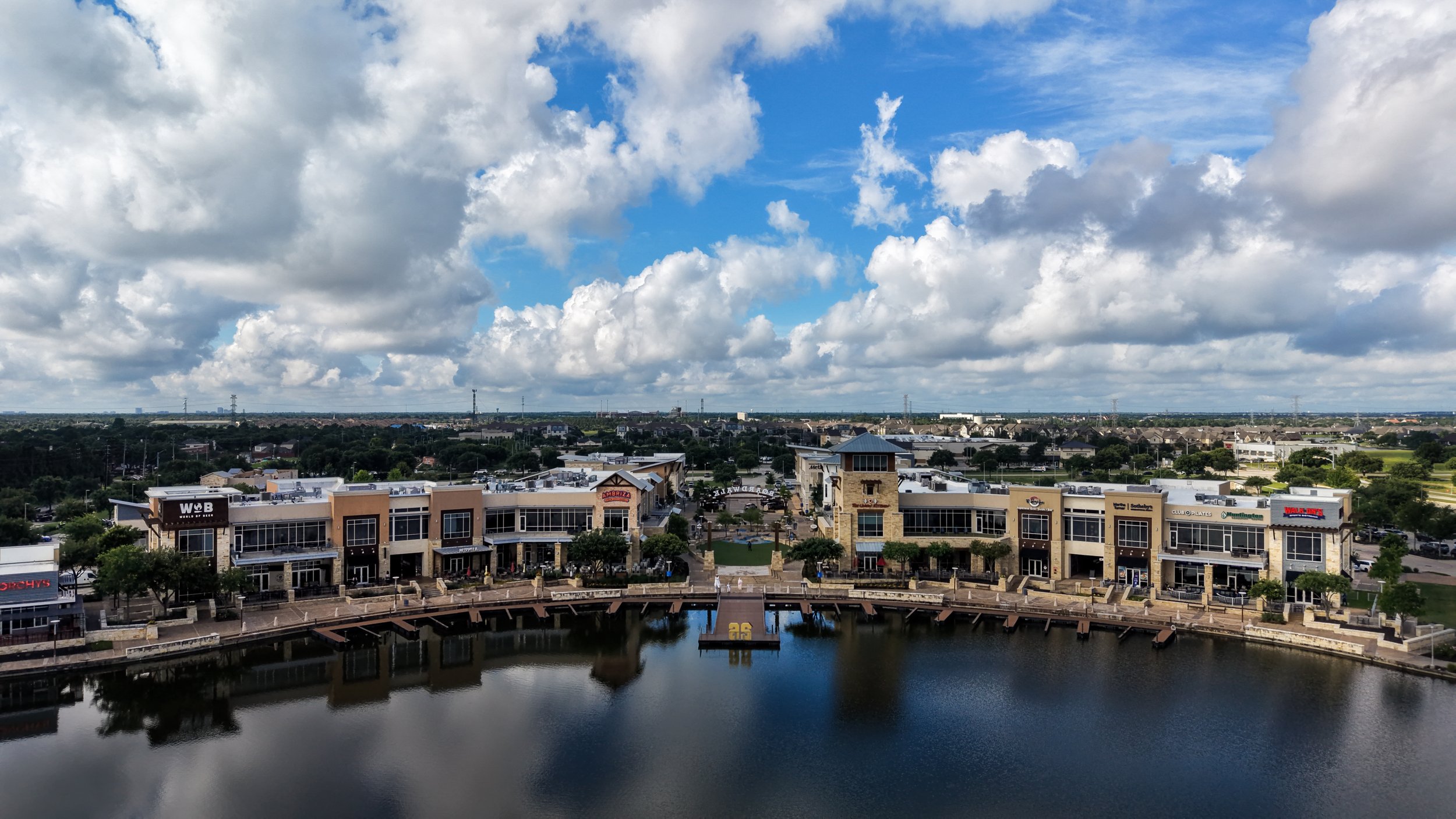 A panoramic view of a modern shopping center with various storefronts, a water feature in the foreground, and a cityscape beyond under a partly cloudy blue sky.