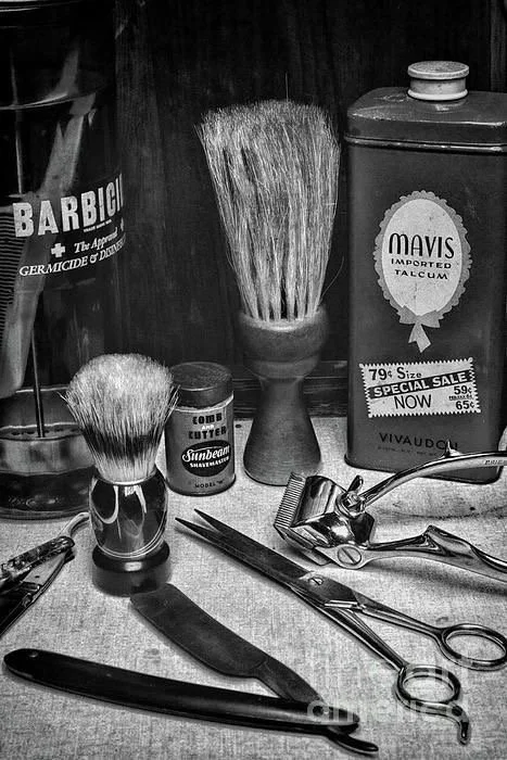 A collection of vintage barber tools and supplies, including shaving brushes, a straight razor, scissors, a jar labeled 'Sunbeam' shaving cream, and tins of talcum powder, arranged on a wooden surface.