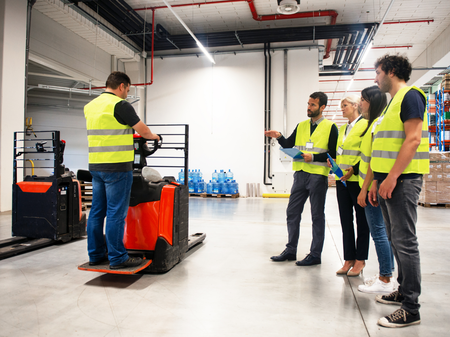 A group of five people in safety vests standing in a warehouse, observing a worker operating a forklift. The warehouse has shelves with boxes and water bottles, with exposed piping and a concrete floor.