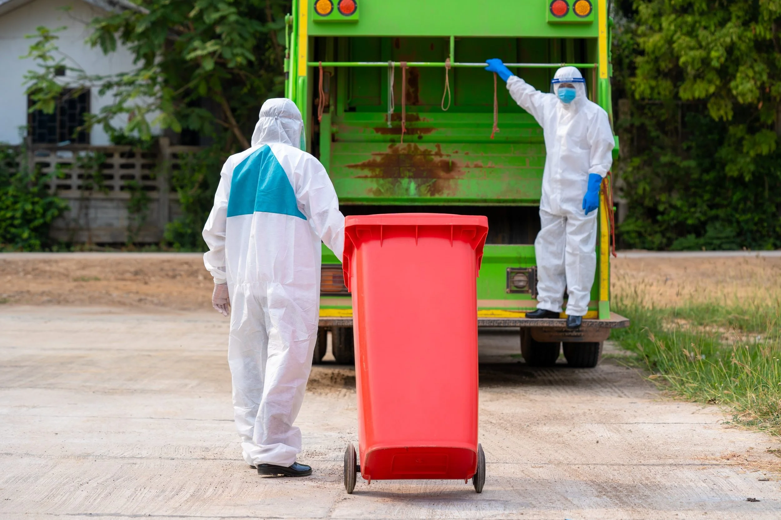 Two people in full protective hazmat suits and masks, working near a green trash truck on a concrete driveway. One person is pushing a large red trash bin, while the other is on the truck, possibly instructing or inspecting.
