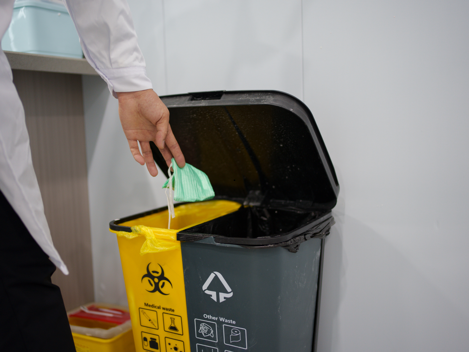 A person wearing a white coat disposing of medical waste in a yellow biohazard trash bin in a medical facility.