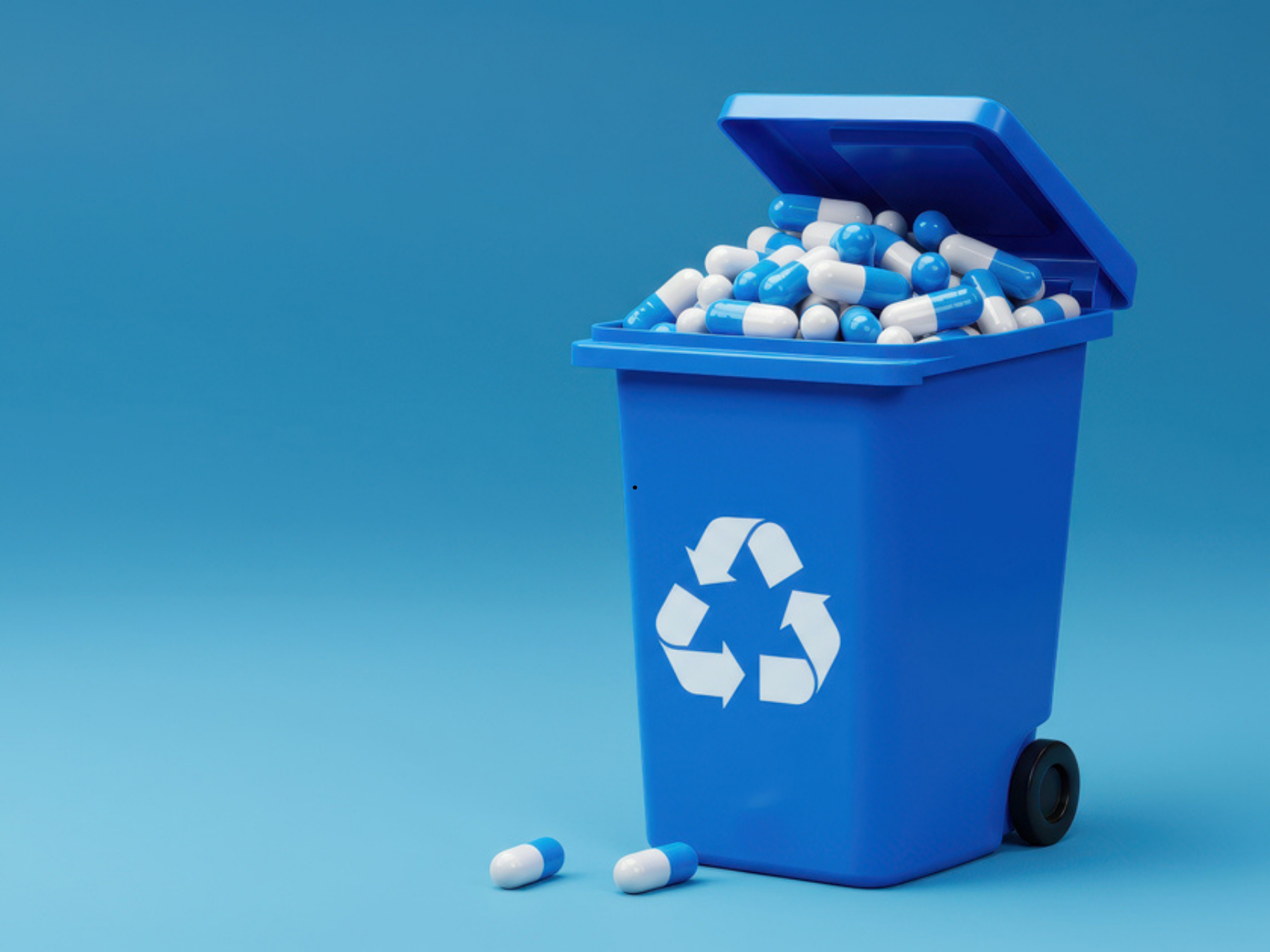 Blue recycling bin filled with white and blue capsules, with some capsules spilled on the blue surface in front. The bin has a recycling symbol on it.