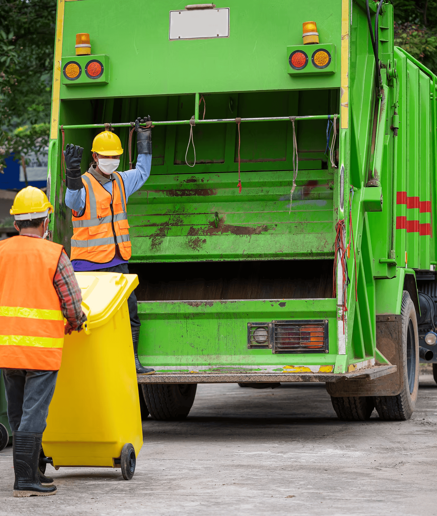 Two sanitation workers, wearing orange safety vests and yellow helmets, are loading trash into a green garbage truck, with one worker holding a yellow trash bin and the other helping to lift it.