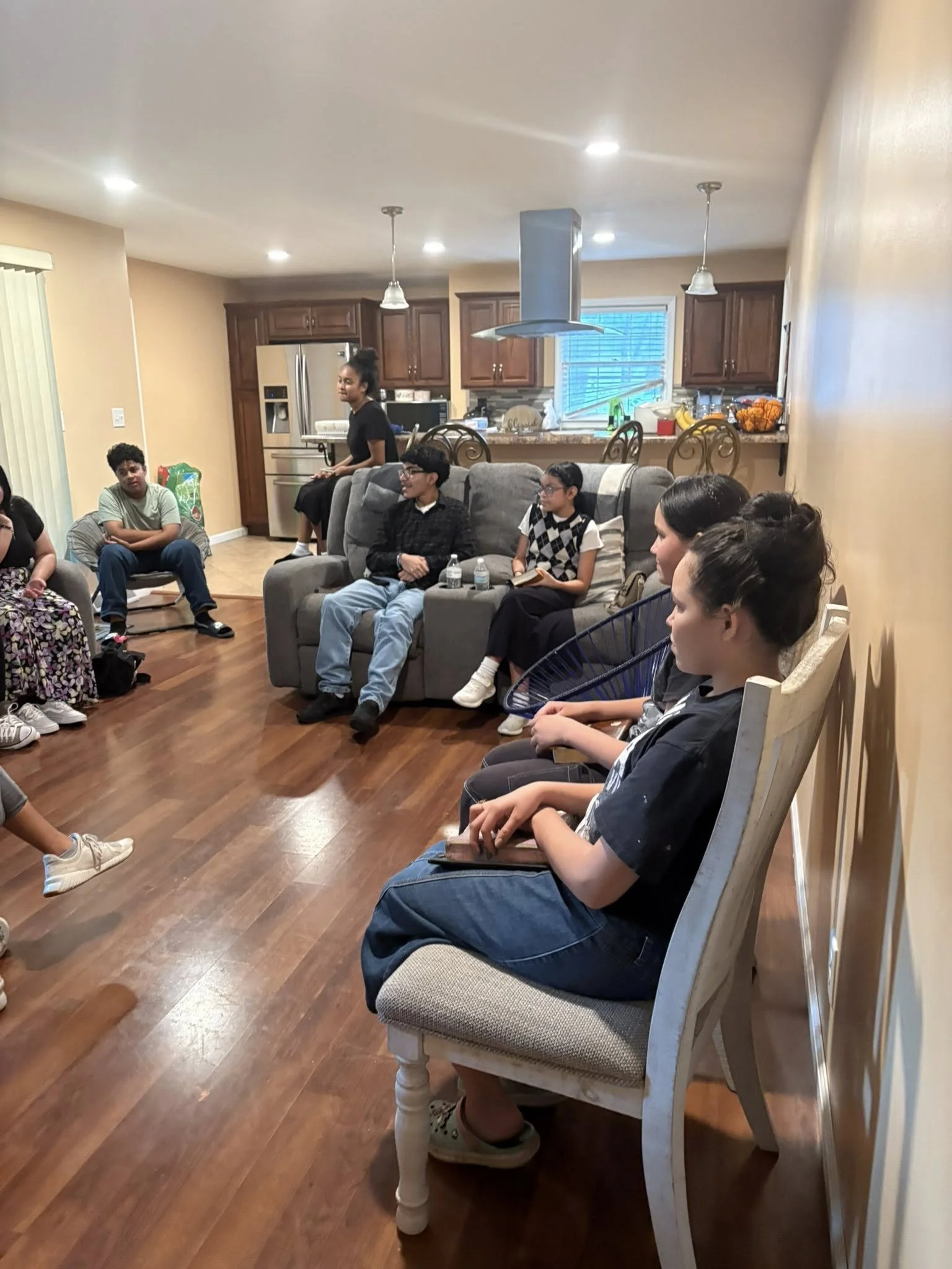 Grupo de jóvenes y adultos sentados en un salón de una casa, participando en una reunión o discusión, con cocina visible al fondo.