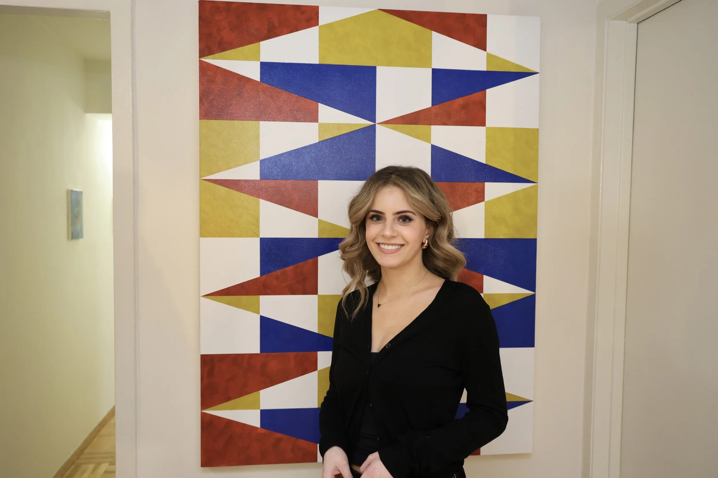 A woman with wavy blonde hair smiling, dressed in a black top with a V-neck, standing in front of a colorful geometric abstract art piece on a white wall. The art features red, yellow, blue, and white triangles and parallelograms arranged in a symmetrical pattern.