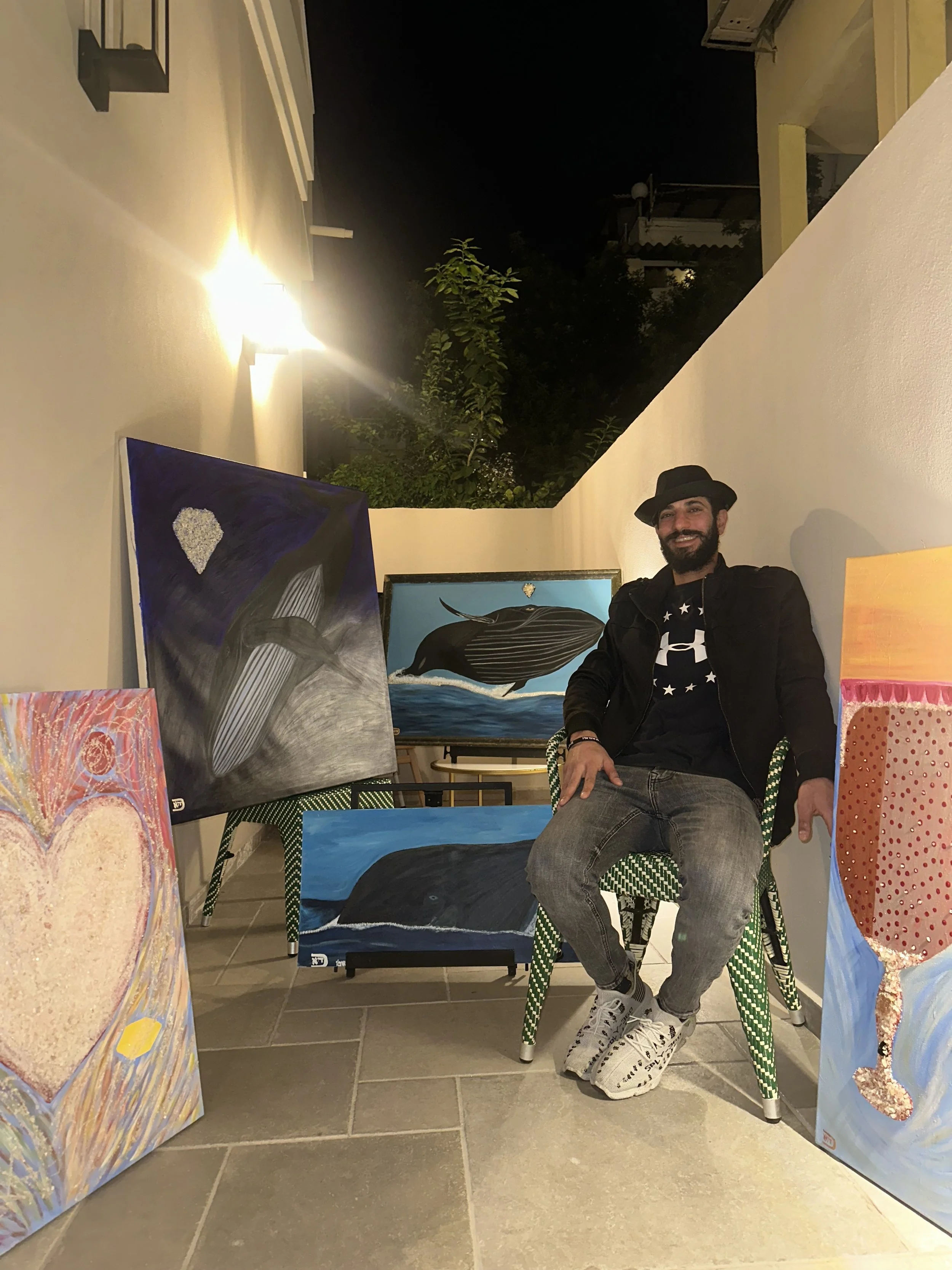 A man sitting on a green and white checkered chair in an art gallery, surrounded by colorful paintings, with a white wall and night sky in the background.