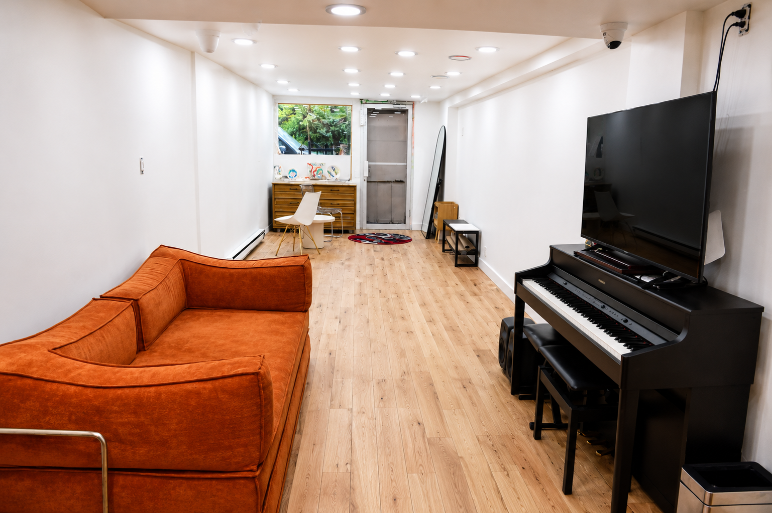 Living room with orange couch, flat-screen TV on a black TV stand, digital piano with a bench, and small table with a mirror, wooden floors, white walls, ceiling lights, and a door leading outside.