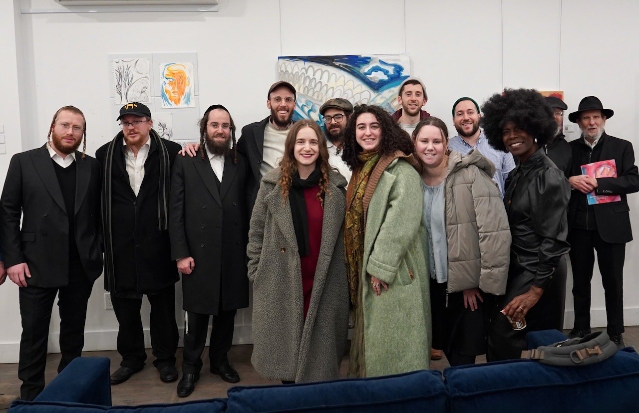 Group of people standing together indoors, smiling, at an art exhibition or gallery. Some are dressed in formal or stylish clothing, and recent artwork is visible on the wall behind.