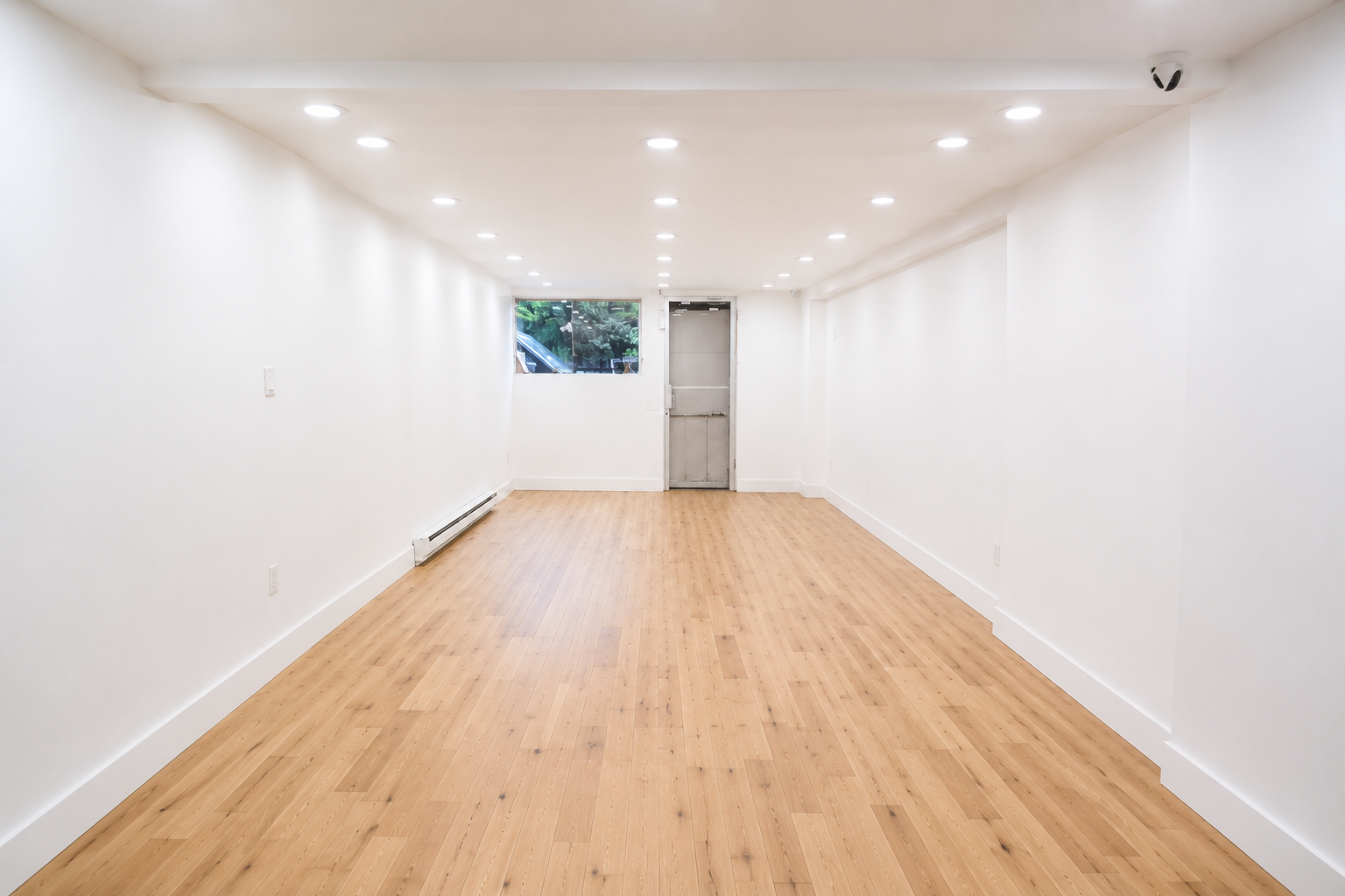 Empty room with white walls, wooden floor, ceiling lights, a small window, and a metal door at the far end.