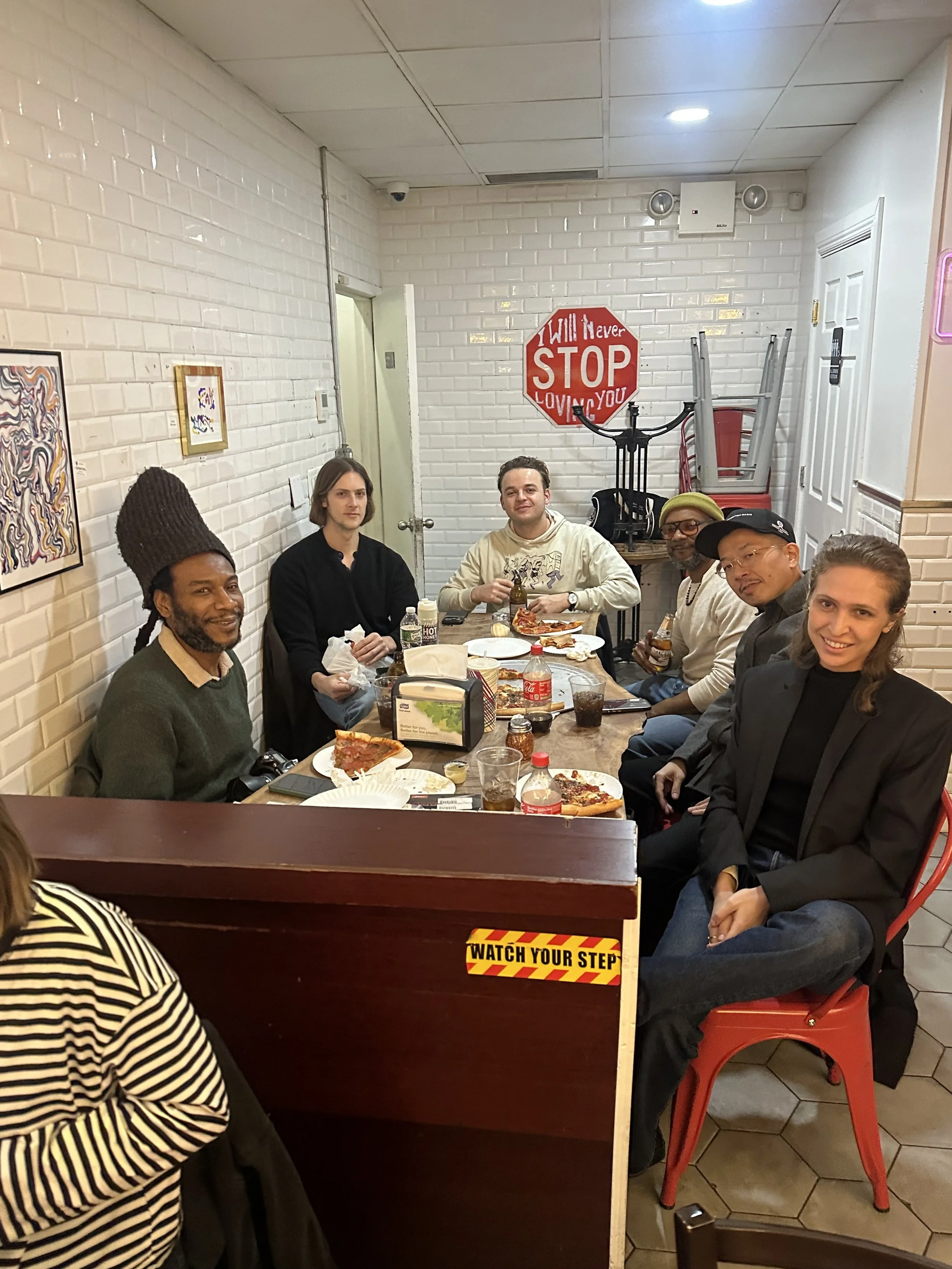 A group of seven diverse people seated at a dining table in a restaurant, enjoying pizza, drinks, and smiling. The restaurant has white brick walls, framed artwork, a surveillance camera, a red stop sign with the message 'I Will Never STOP LOVING YOU,' and various chairs and tables.