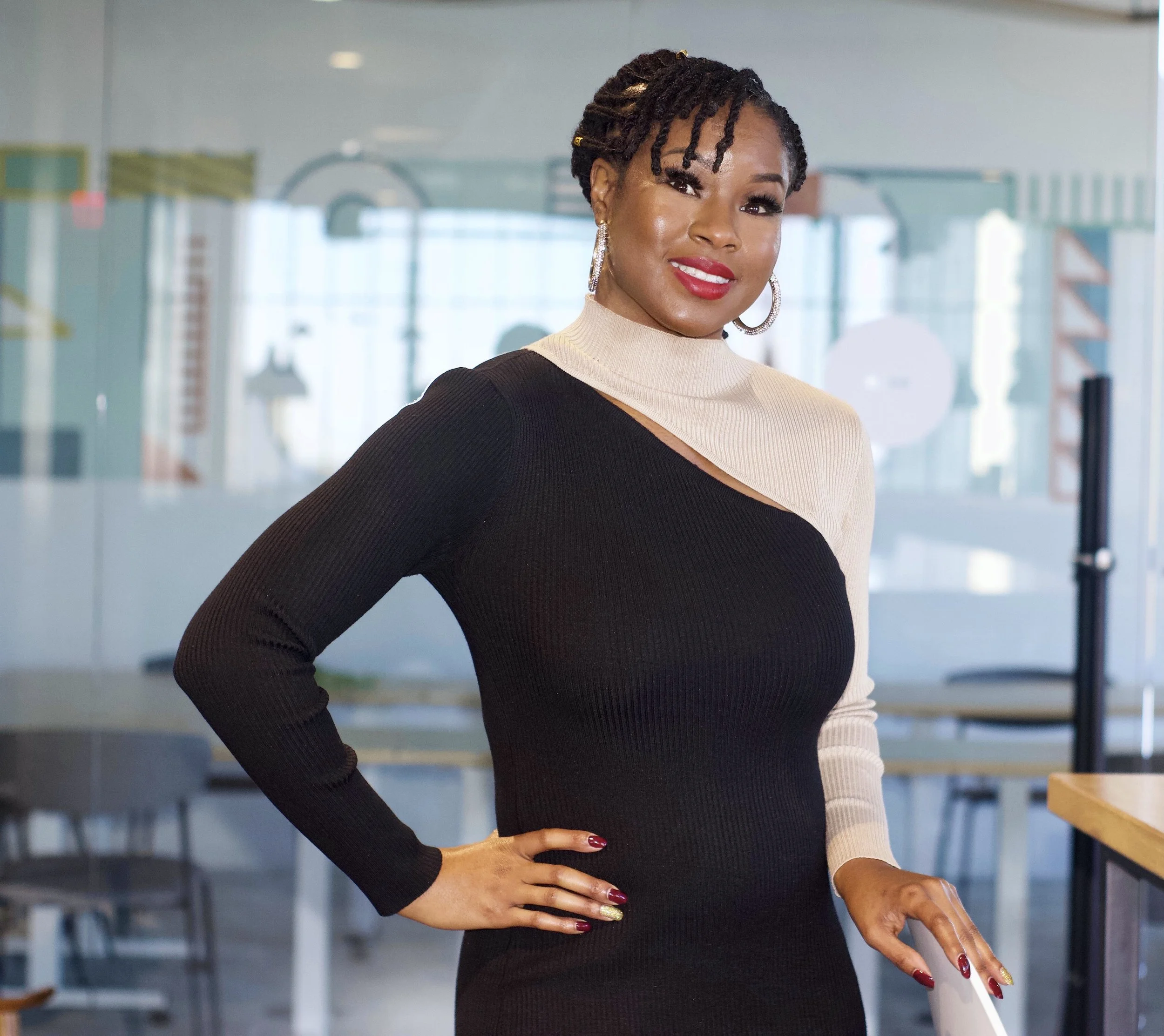 A woman with dark skin and dreadlocks styled back, wearing a black and beige color-blocked, long-sleeve knit dress, standing in a modern, glass-enclosed office space, Book Coach