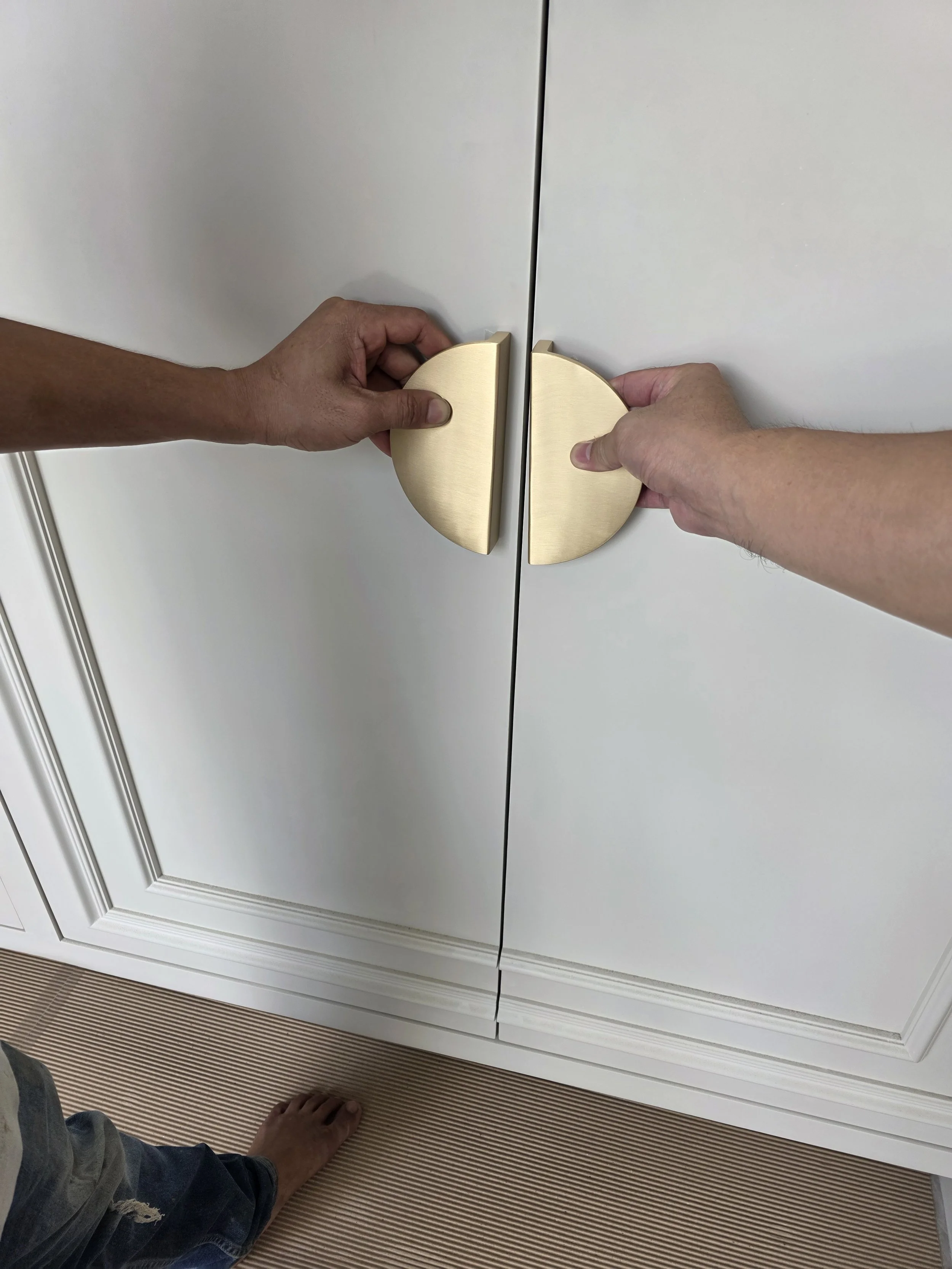 Two people are opening a white cabinet with gold round handles. One person is pulling on each handle, revealing the cabinet's interior.