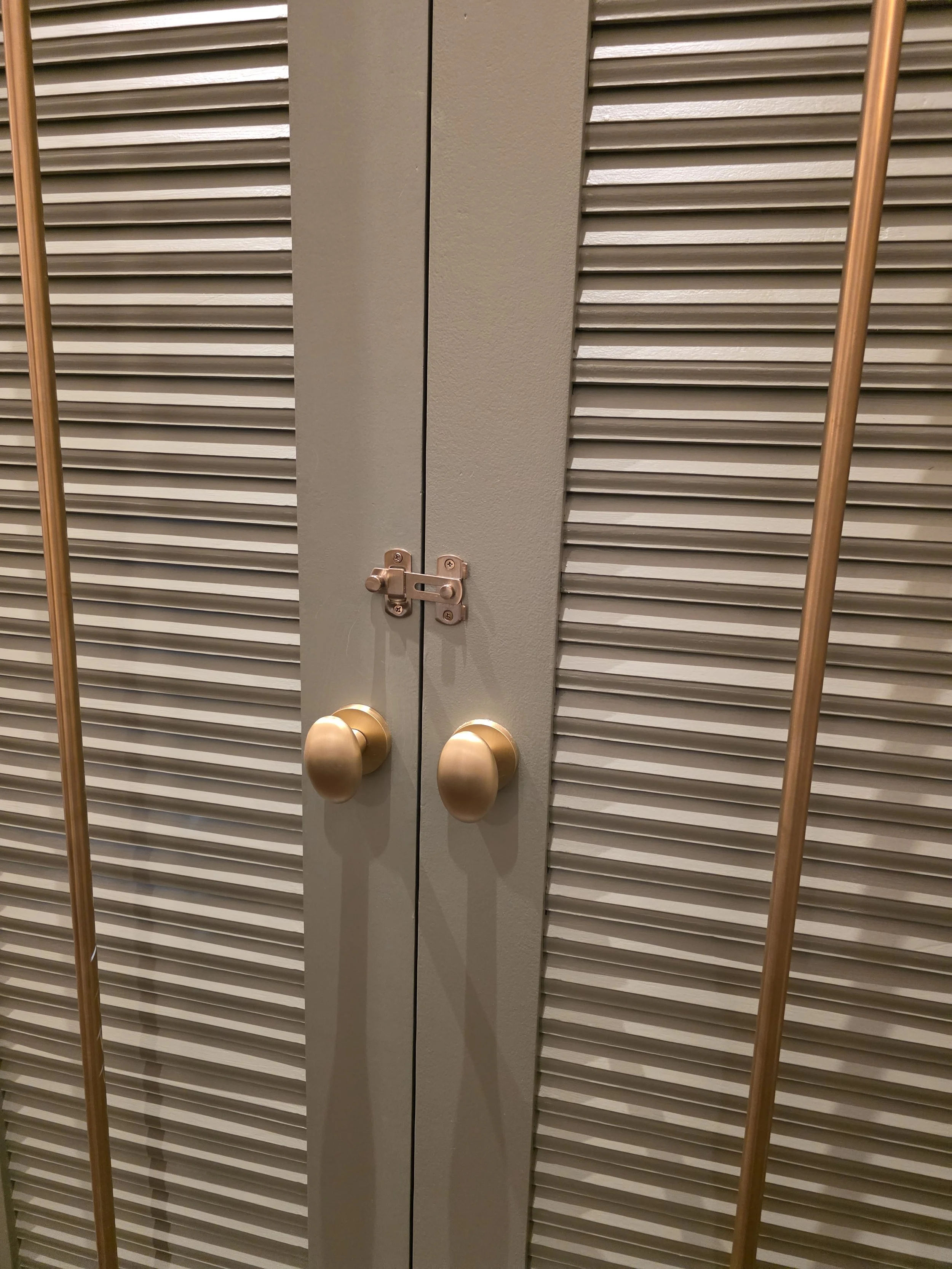 Closet door with gold door knobs and a top latch, surrounded by louvered closet doors with brown vertical trim.