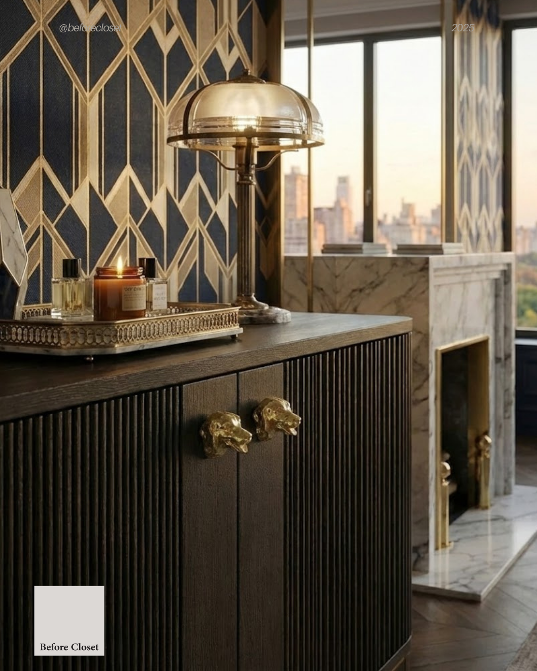 Interior of a stylish room with a wooden cabinet featuring golden animal head handles, a marble fireplace, a large window showing a cityscape at sunset, a decorative tray with candles and perfume bottles, and a vintage table lamp.