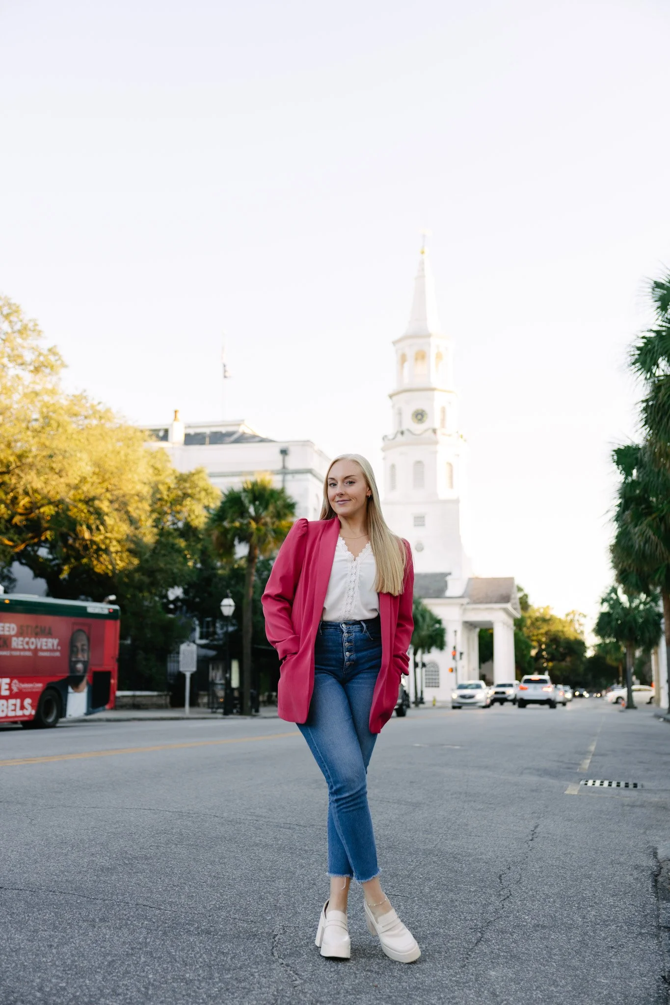A young woman wearing a pink blazer, white blouse, blue jeans, and white platform shoes standing on a city street with a historic white church in the background, trees with autumn foliage, and cars in the distance.
