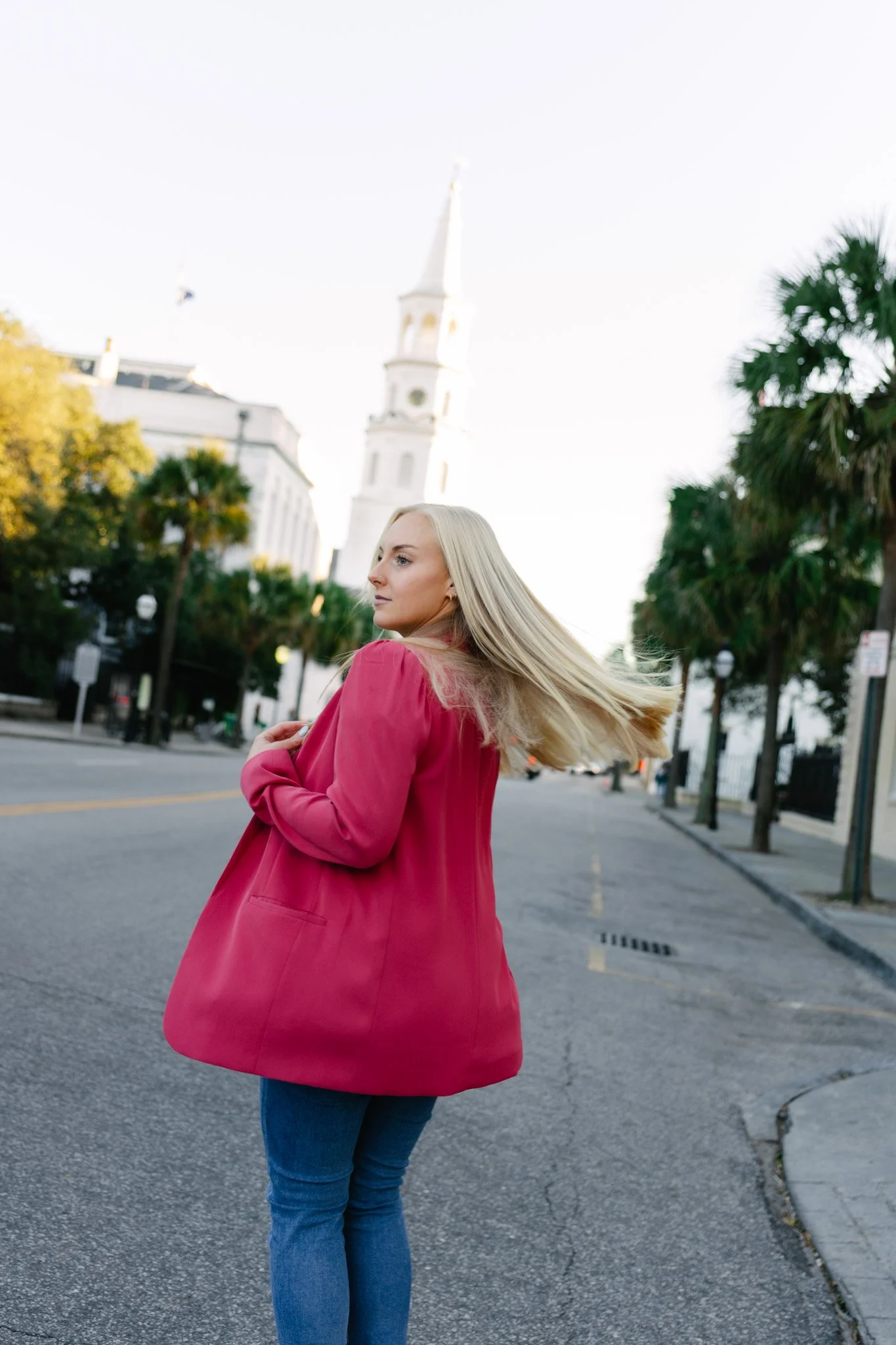 A woman with long blond hair wearing a pink coat and blue jeans standing on a city street with palm trees and a white church steeple in the background.