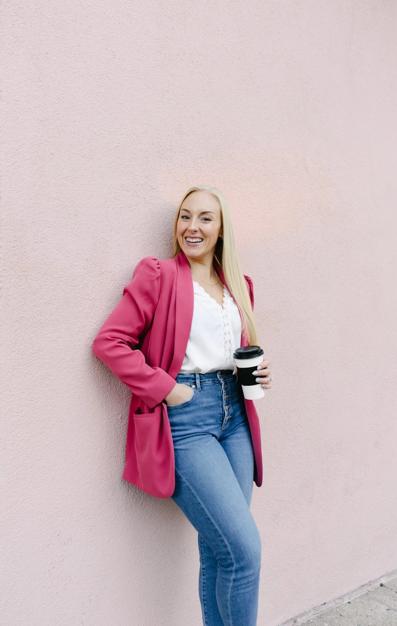 A young woman with long blonde hair, wearing a pink blazer and blue jeans, leaning against a pink textured wall while holding a coffee cup and smiling.