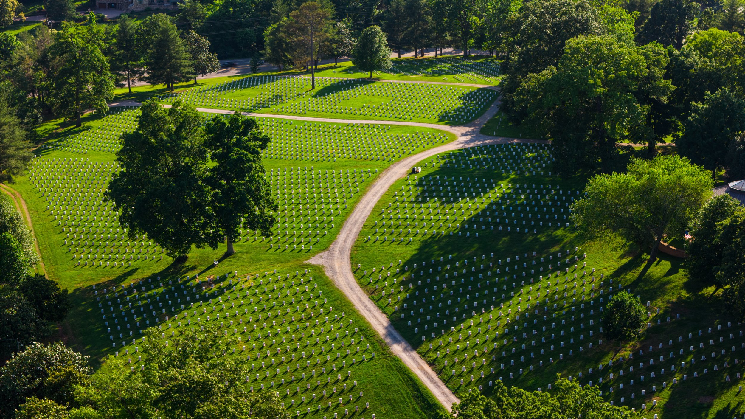 Aerial view of a cemetery with white headstones, surrounded by green trees and grass, with a winding dirt road passing through.