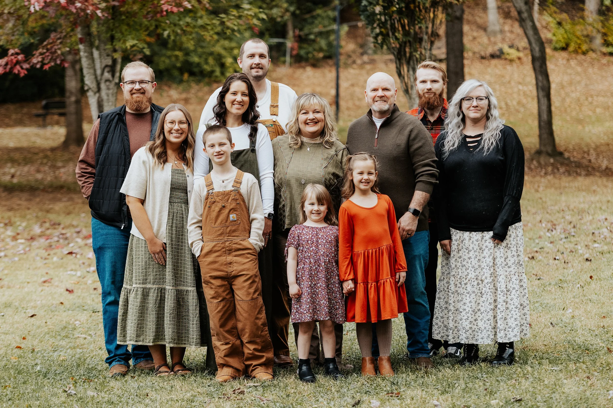 A large family group posing outdoors in autumn, standing on grass with fall trees and fallen leaves in the background.
