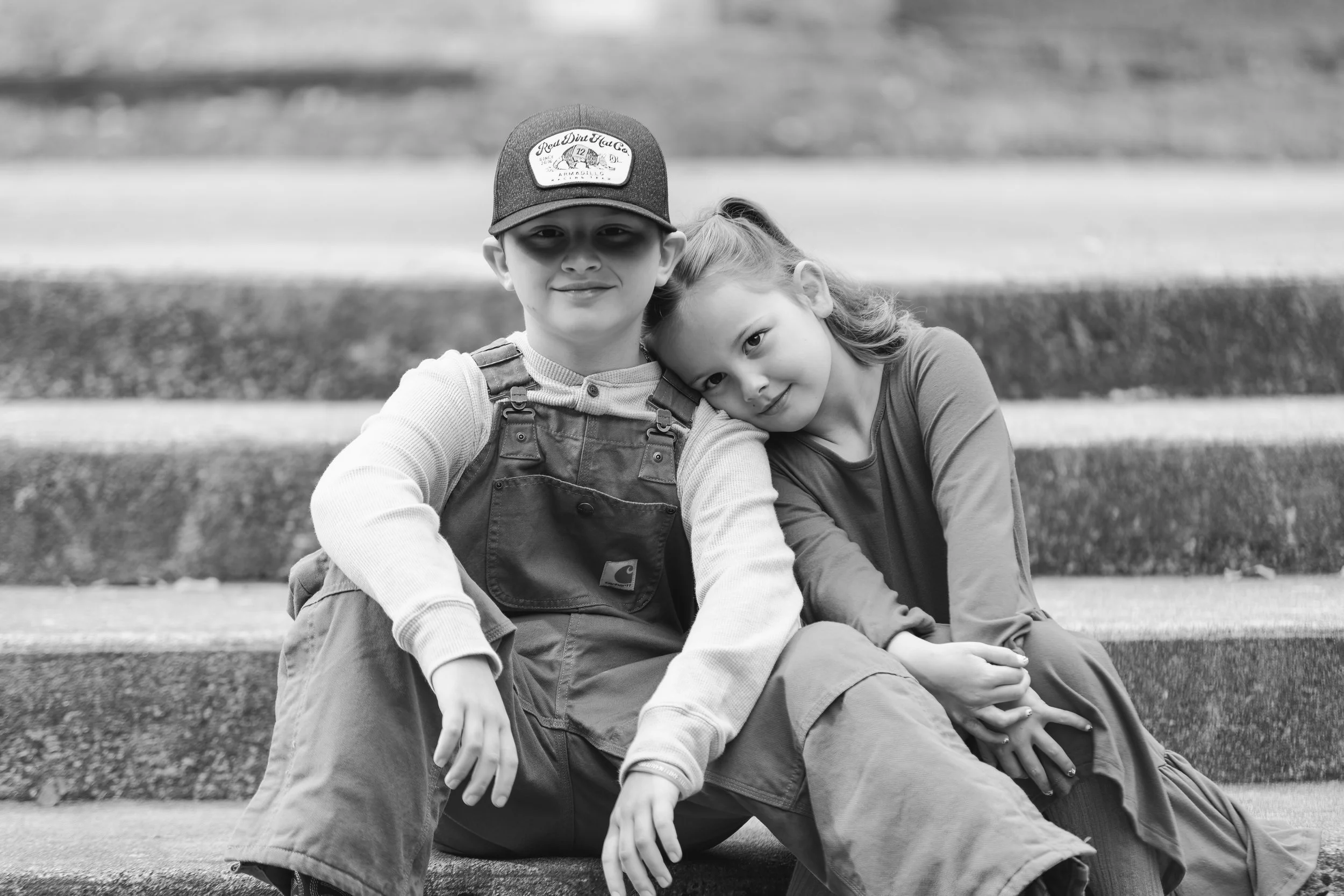 A boy wearing a cap and overalls sitting on steps with a girl resting her head on his shoulder