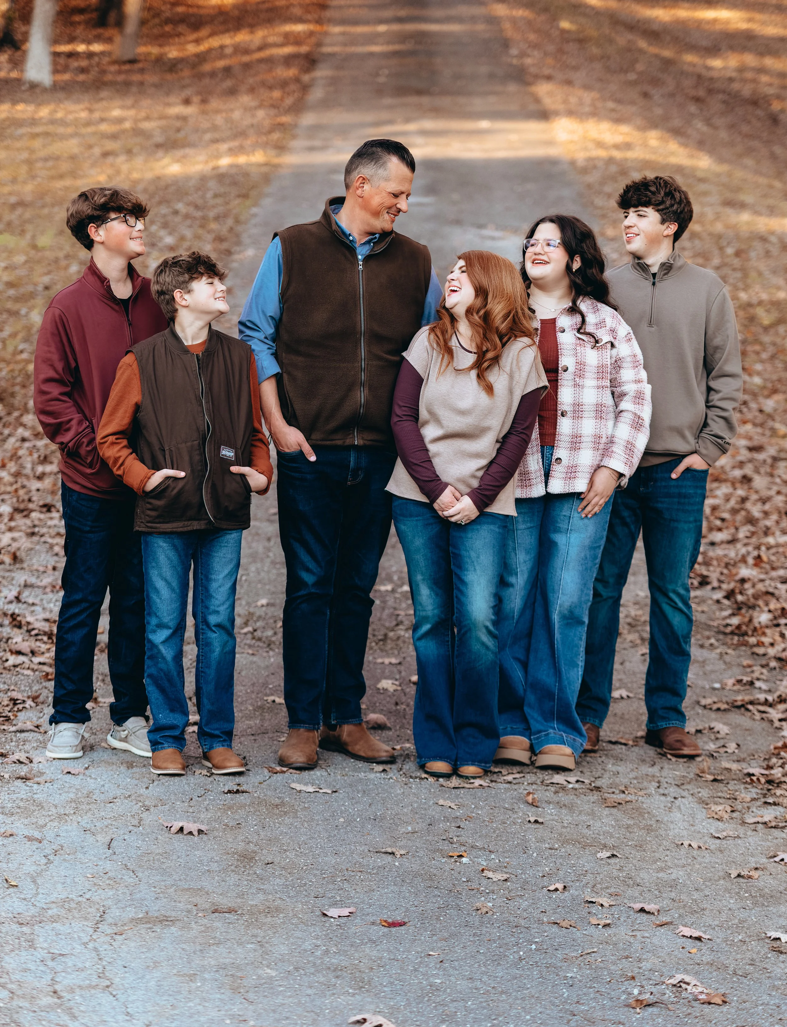 A family of six, including one adult male and five children, standing on a wooded path outdoors during autumn, smiling and looking at each other.