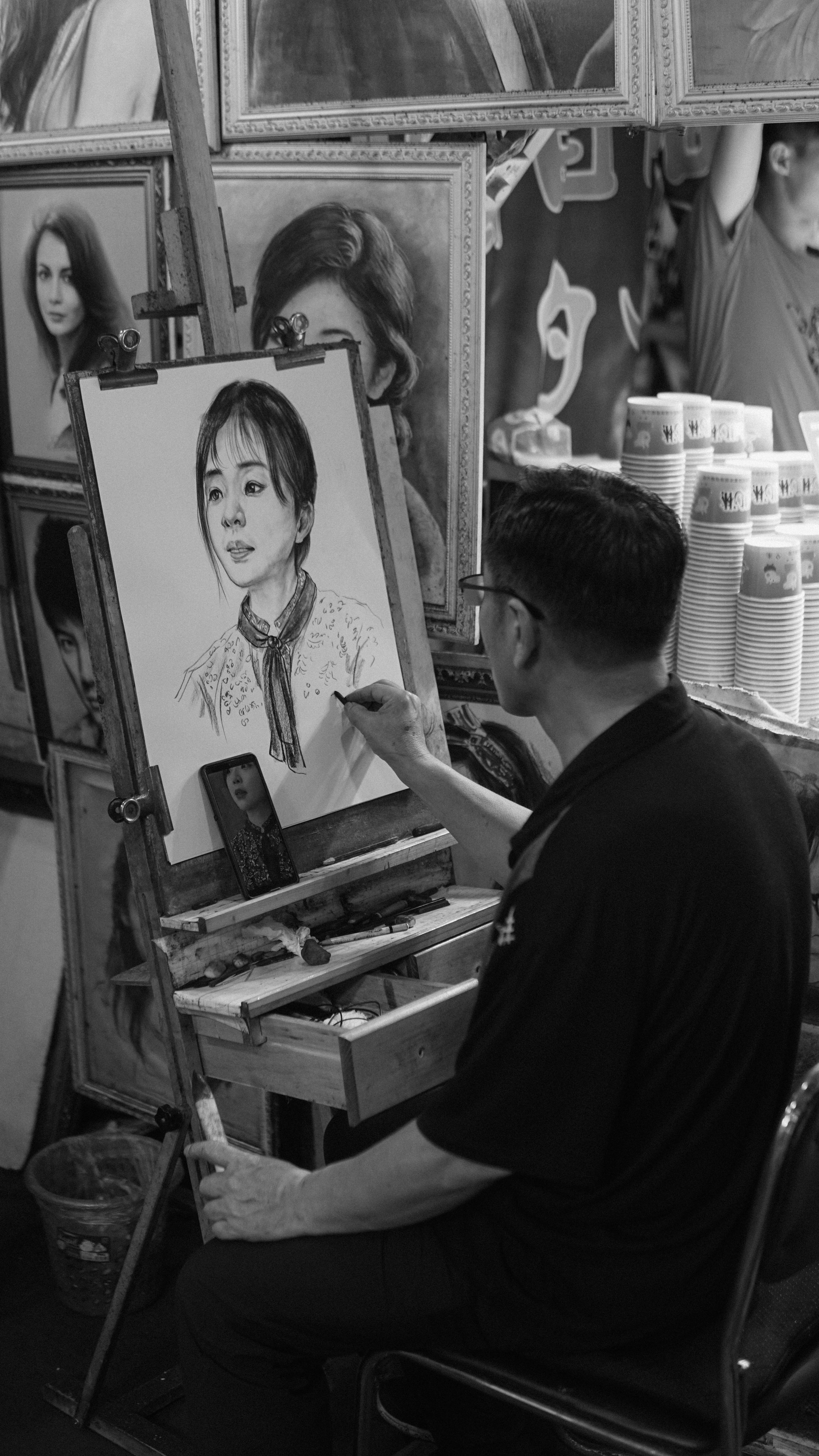 An artist sketching a portrait of a woman in a black shirt and glasses, with several framed portraits hanging behind him at an art fair or exhibition.