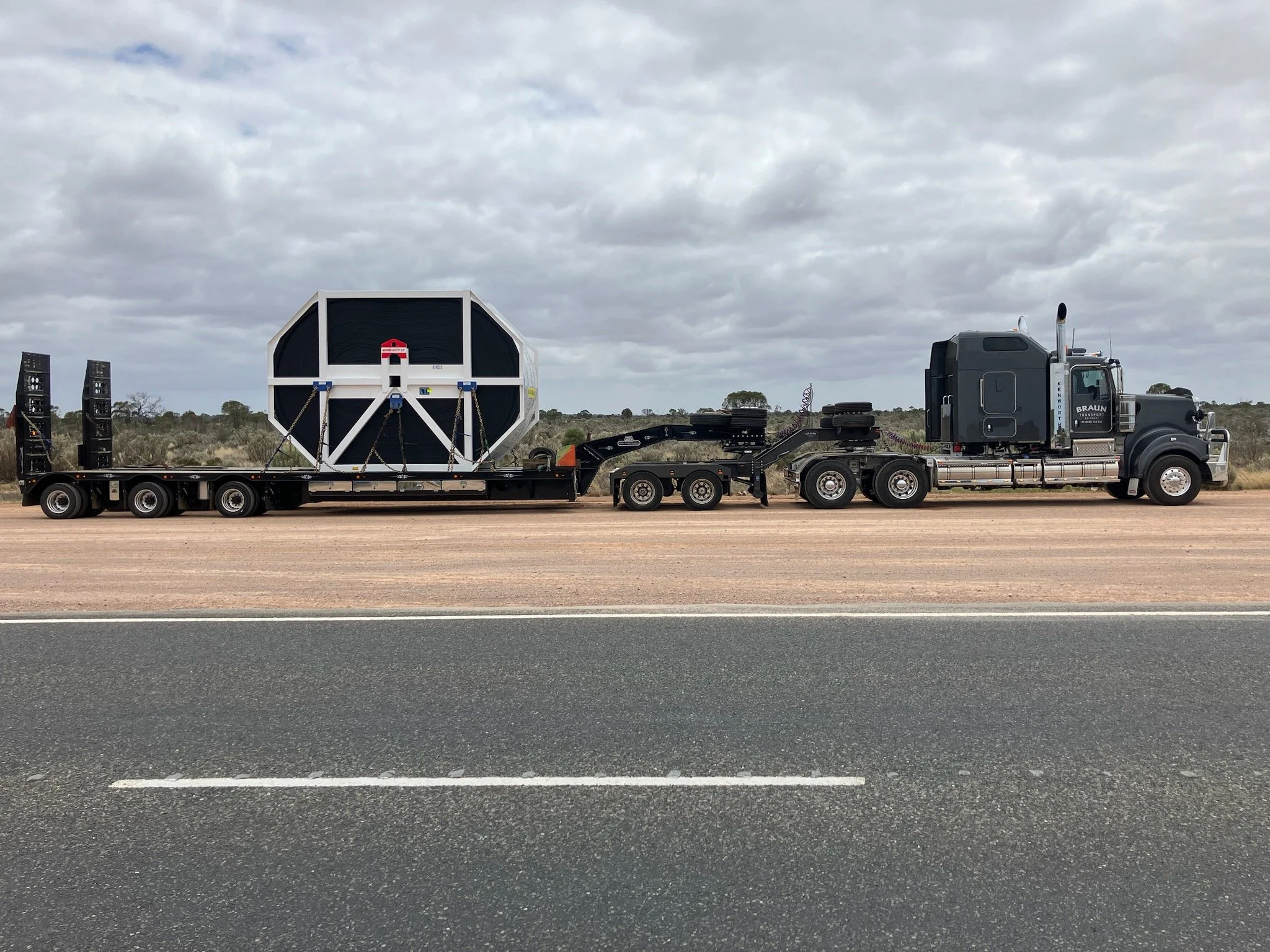 A large semi-truck with a flatbed trailer transporting a large, octagonal, black and white machine or structure on a road with a cloudy sky overhead.
