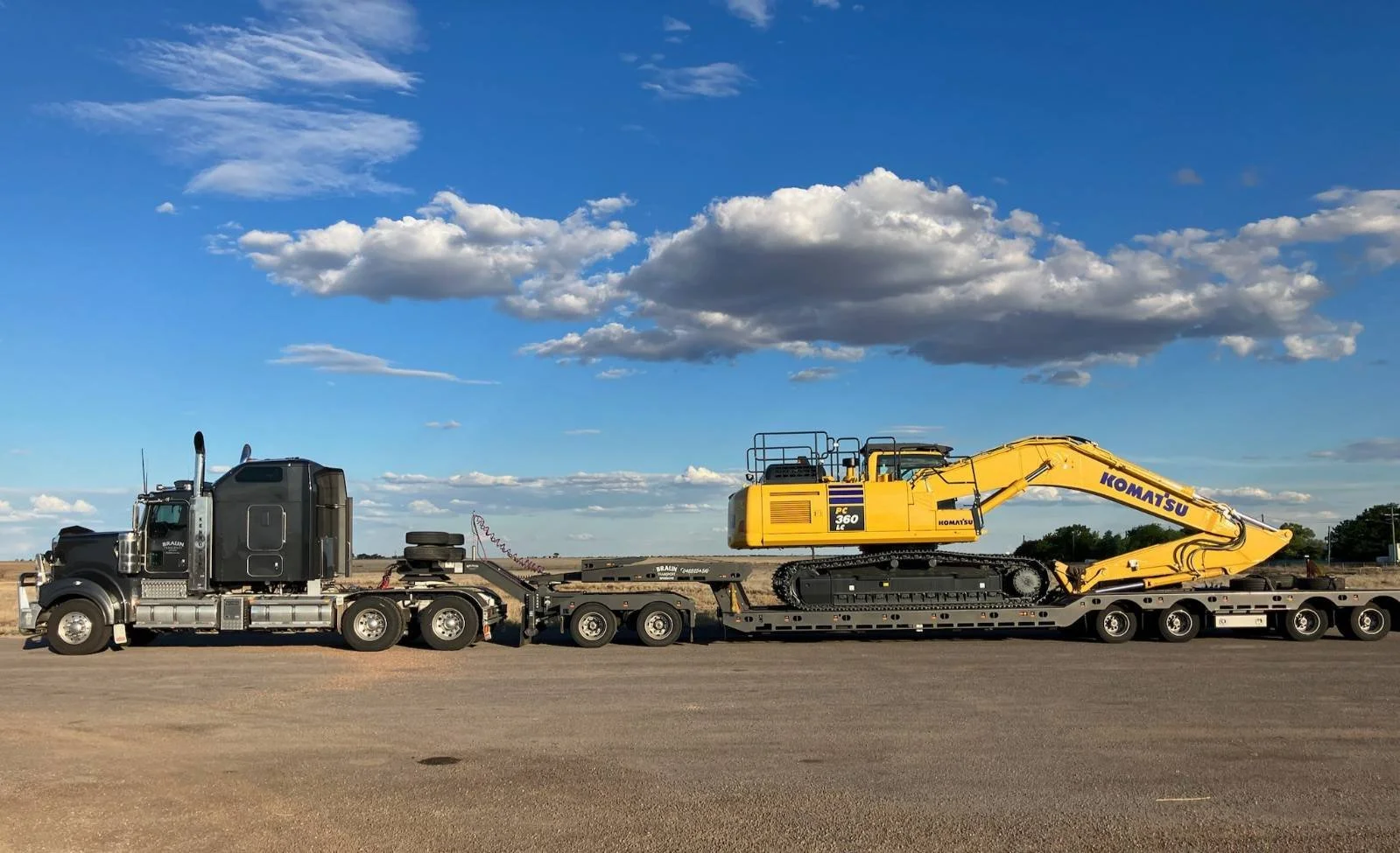 A semi-truck transporting a yellow Komatsu excavator on a flatbed trailer on an open road under a blue sky with scattered clouds.