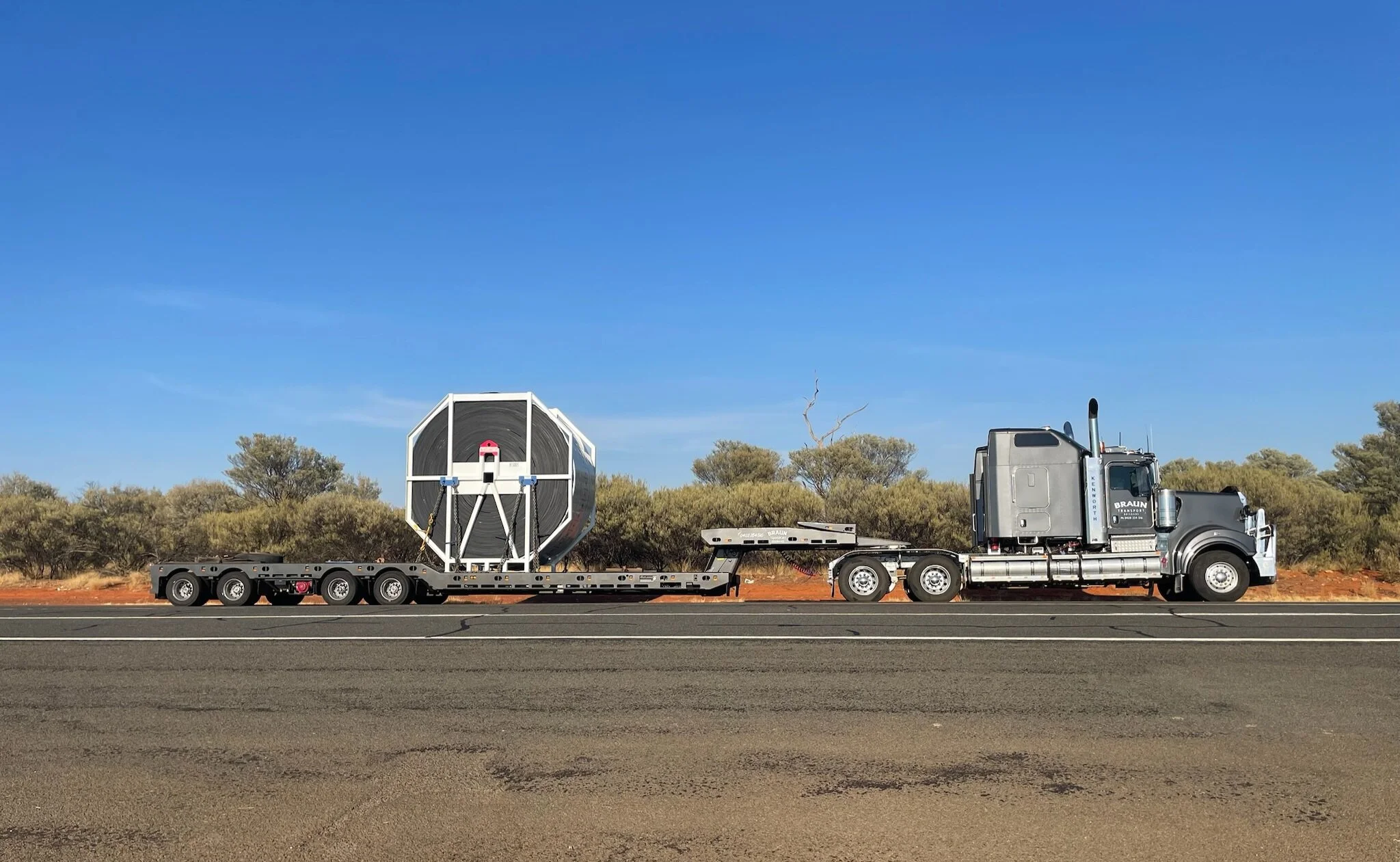 A semi-truck hauling a large, industrial-looking tank on a flatbed trailer on a highway with desert vegetation and a clear blue sky.