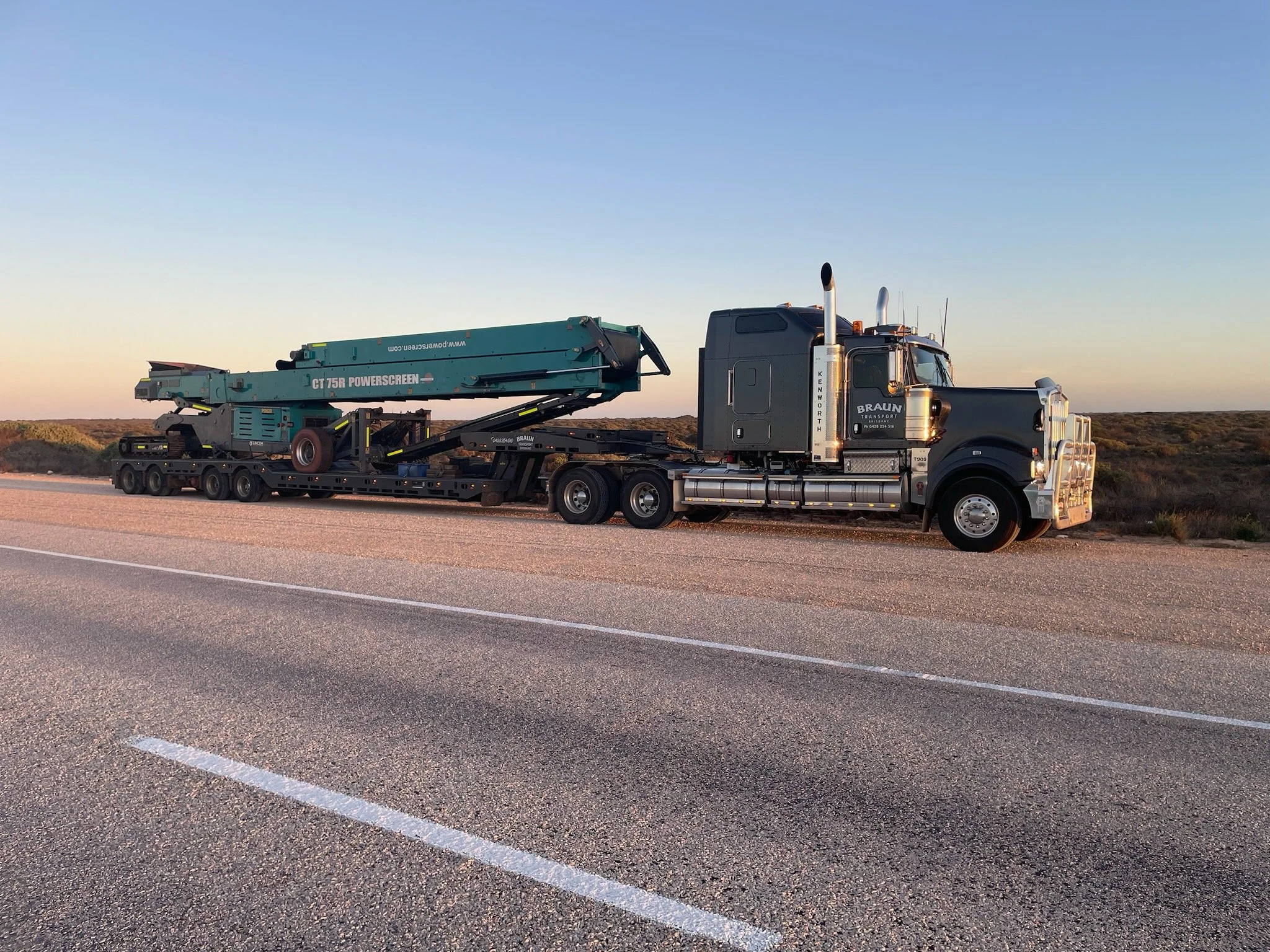 Semi-truck with flatbed trailer carrying a large green piece of equipment on the side of a rural road at sunset.