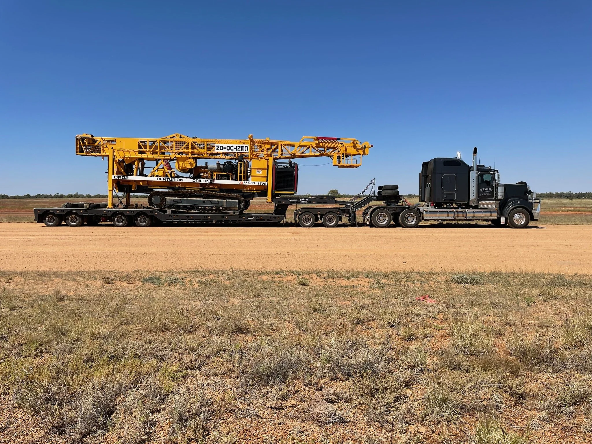 A black semi-truck is parked on a dirt road, carrying a yellow drilling machine on its flatbed trailer, with open fields and a blue sky in the background.