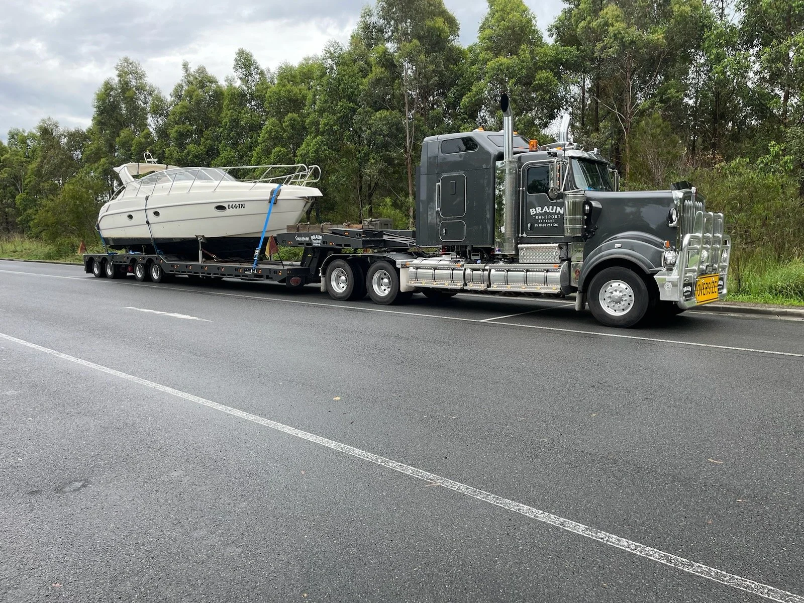 Semi-truck transporting a white boat on a highway surrounded by trees.