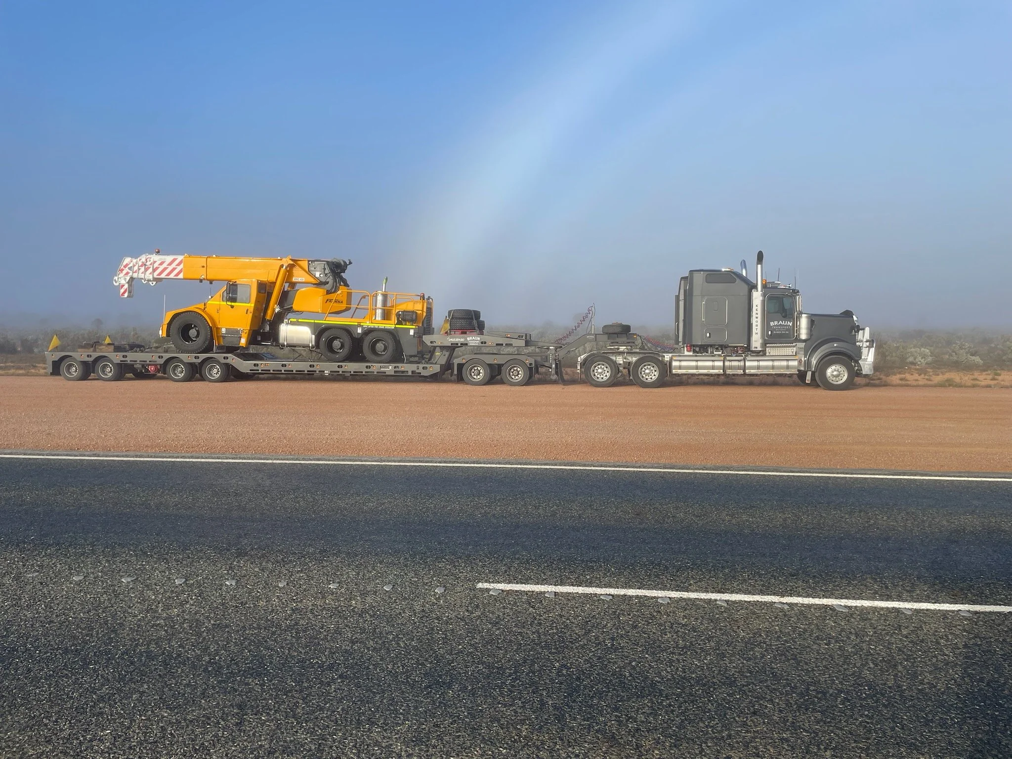 A semi-truck hauling a yellow crane and construction equipment on a flatbed trailer along a highway in a desert landscape.