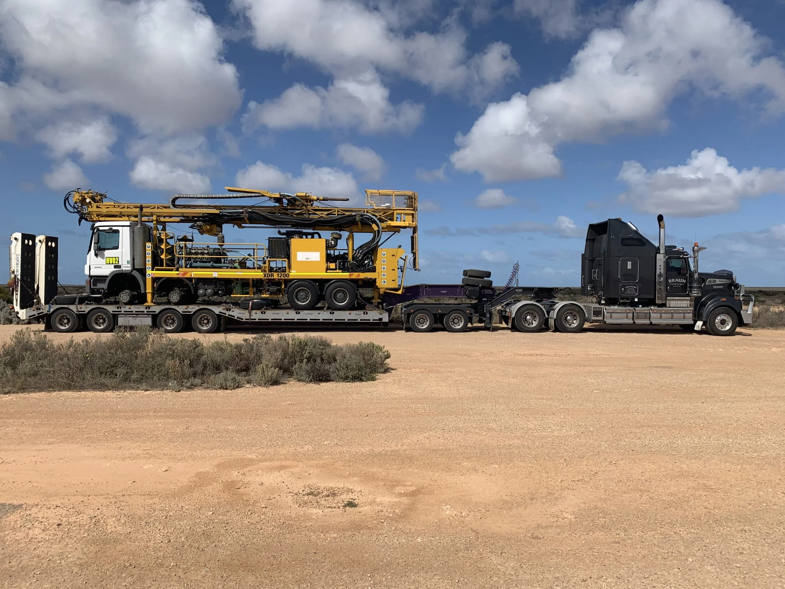 A large black semi-truck hauling industrial equipment across a sandy desert landscape under a partly cloudy sky.
