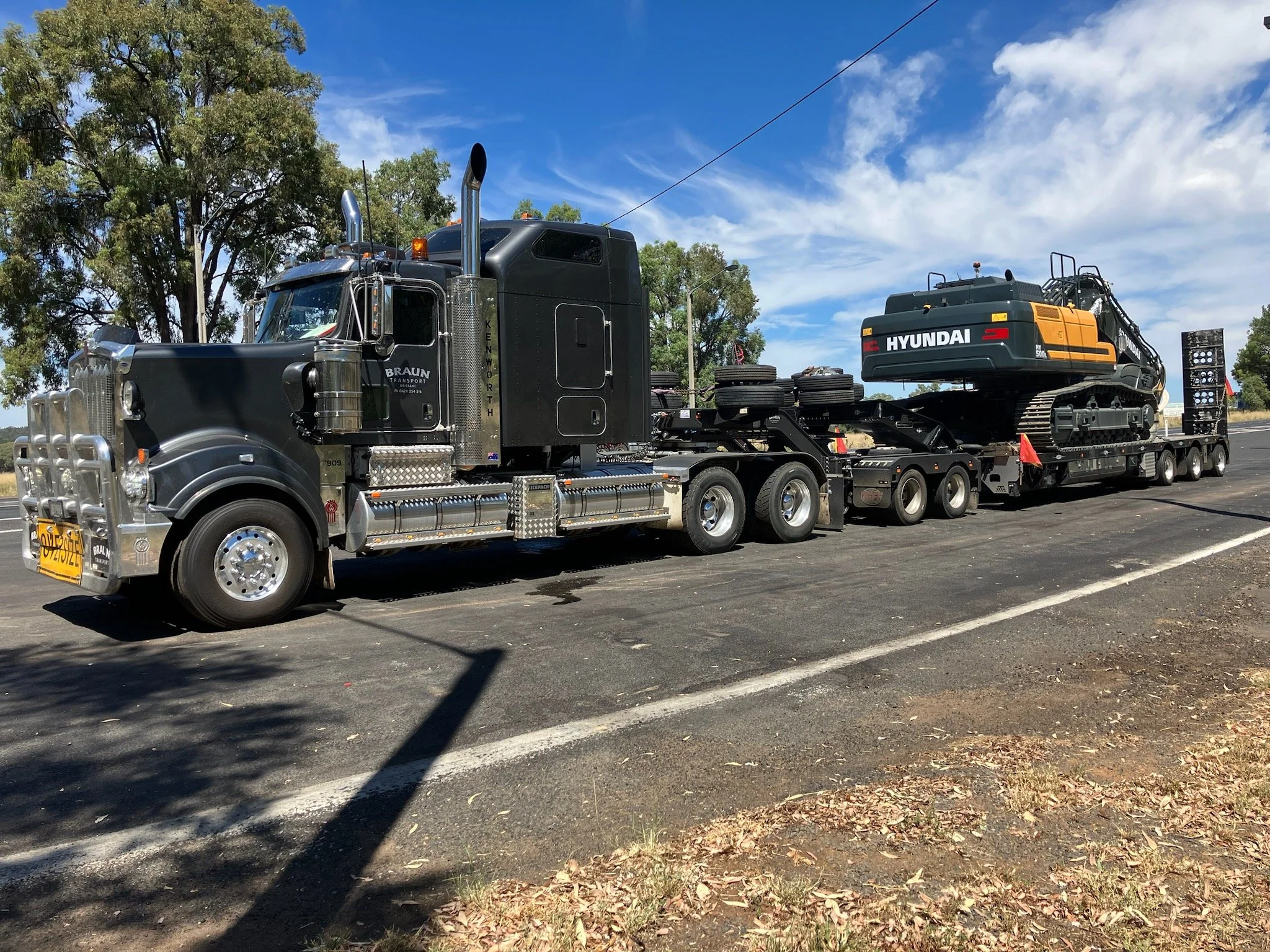 A black semi-truck with chrome details transporting a Hyundai excavator on a flatbed trailer, parked on a road with trees and a blue sky in the background.
