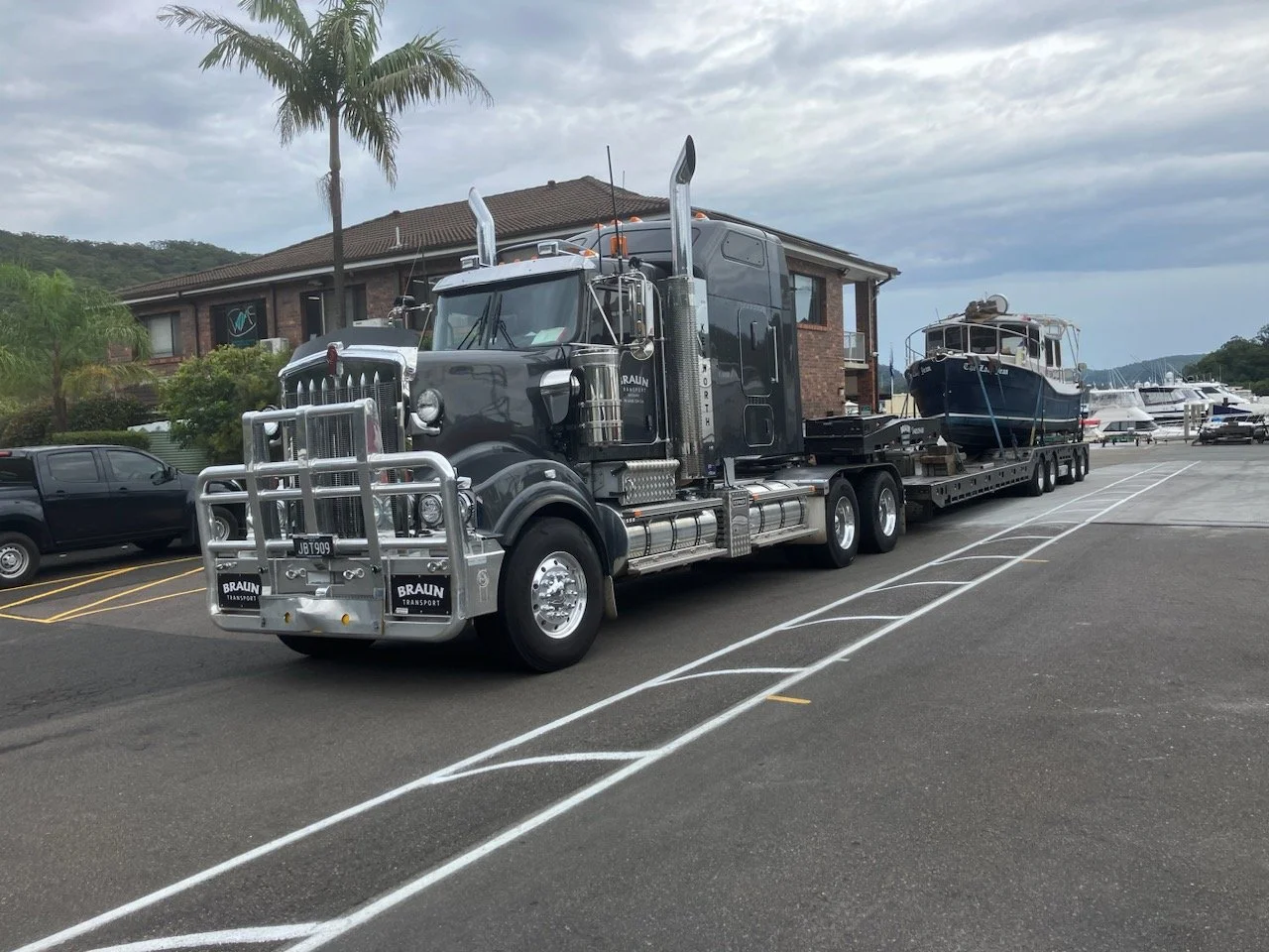 A large black semi-truck transporting a boat on a flatbed trailer parked in a lot with other vehicles, including a black pickup truck, near a brick building and trees, with a cloudy sky overhead.