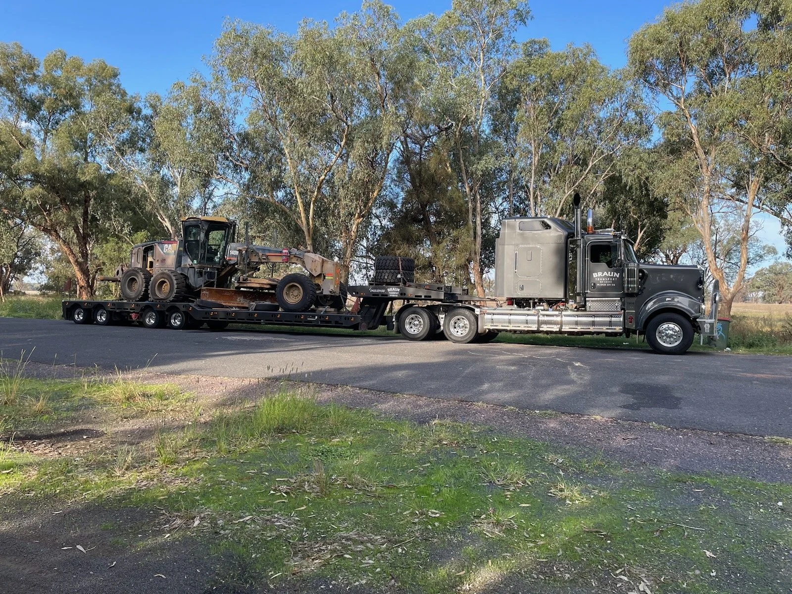 Black semi-truck with a flatbed trailer transporting a vintage bulldozer on a tree-lined road.