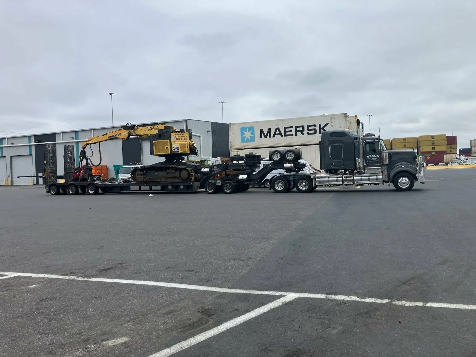 A large semi-truck with a flatbed trailer carrying construction equipment, including a small excavator and a tracked vehicle, parked in a lot with a warehouse and stacked shipping containers in the background.