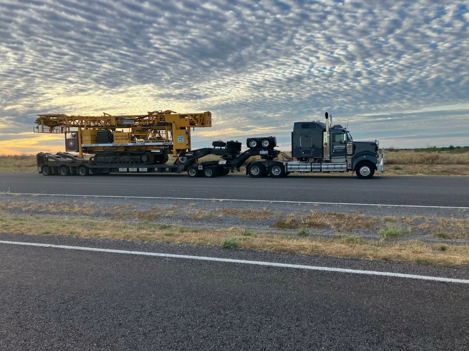 A semi-truck with a flatbed trailer on the highway carrying construction equipment, including a large yellow drilling machine, during sunset with a cloudy sky.