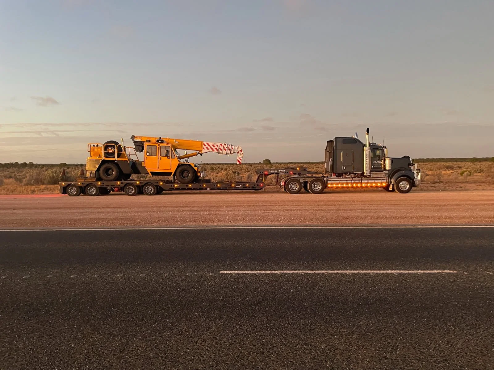 A semi-truck transporting a construction vehicle on a flatbed trailer along a highway at sunset.