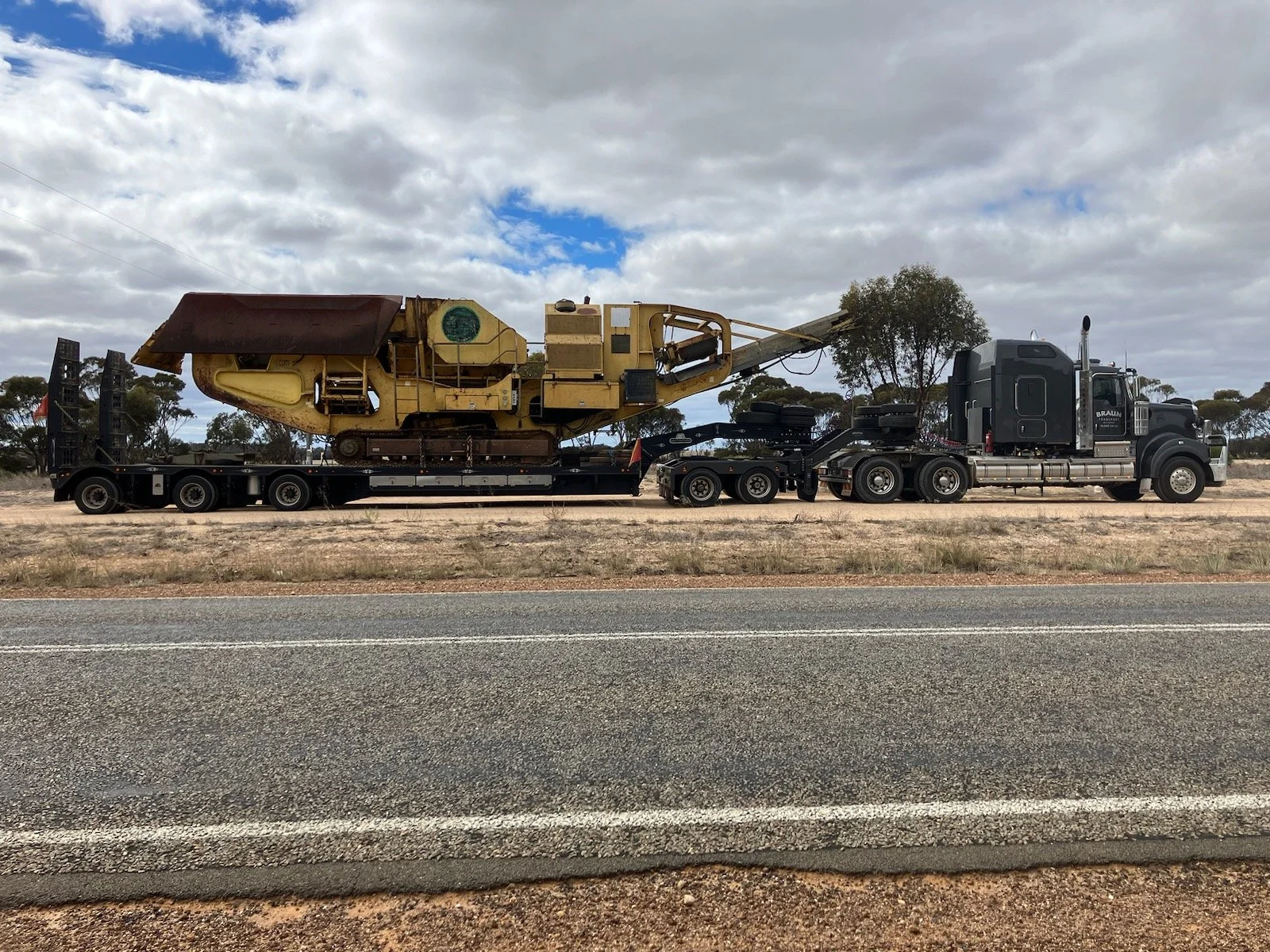 A semi-truck carrying a large, yellow construction machine on a flatbed trailer parked on the side of a road.
