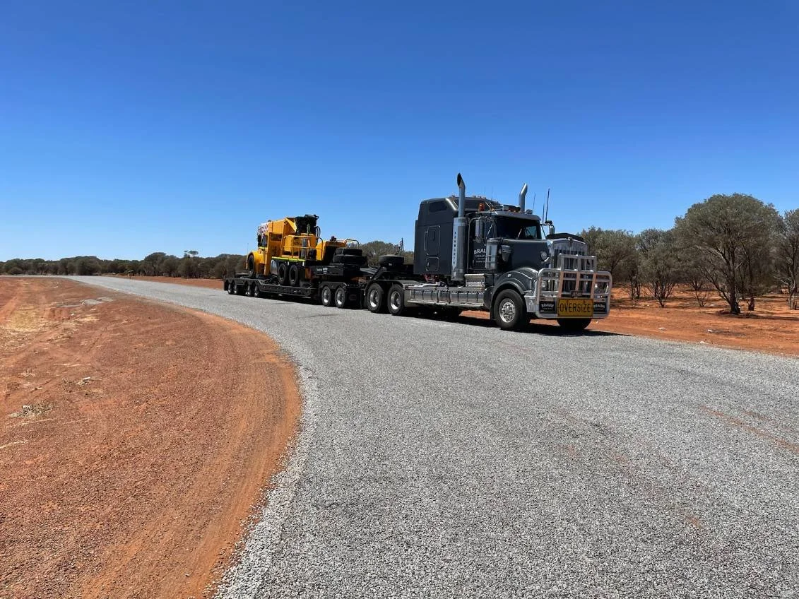 A semi-truck carrying large construction equipment on a gravel road in a dry, sparse landscape with trees in the background and a clear blue sky.