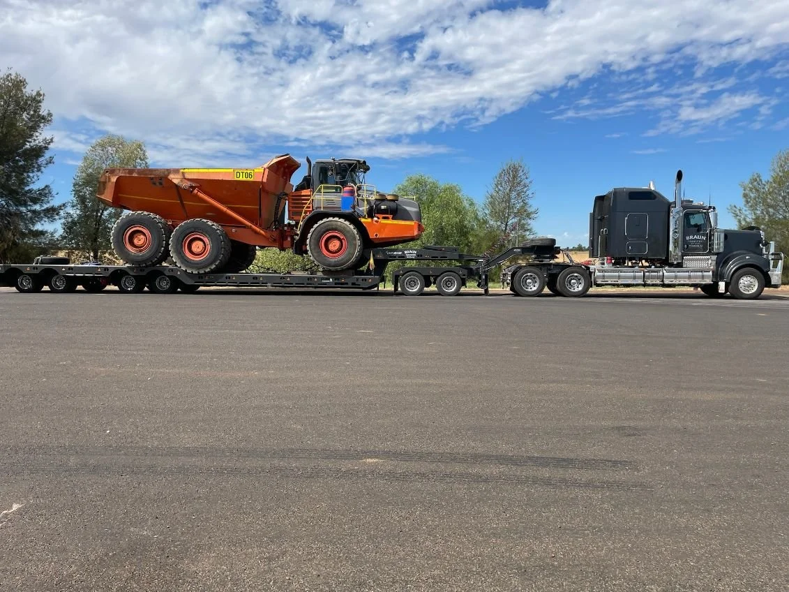A semi truck transporting a large orange construction vehicle on a flatbed trailer on a paved road, with trees and a partly cloudy sky in the background.