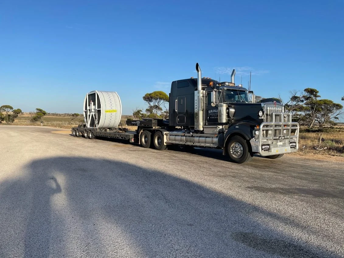 Black semi-truck carrying a large industrial coil in a rural area with trees and clear blue sky