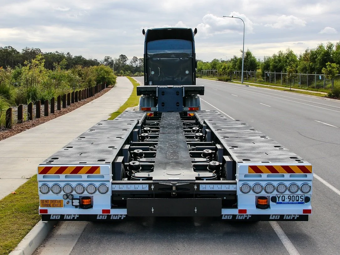 A flatbed truck trailer parked on the side of a road, with a black cab attached and surrounded by green trees and bushes.