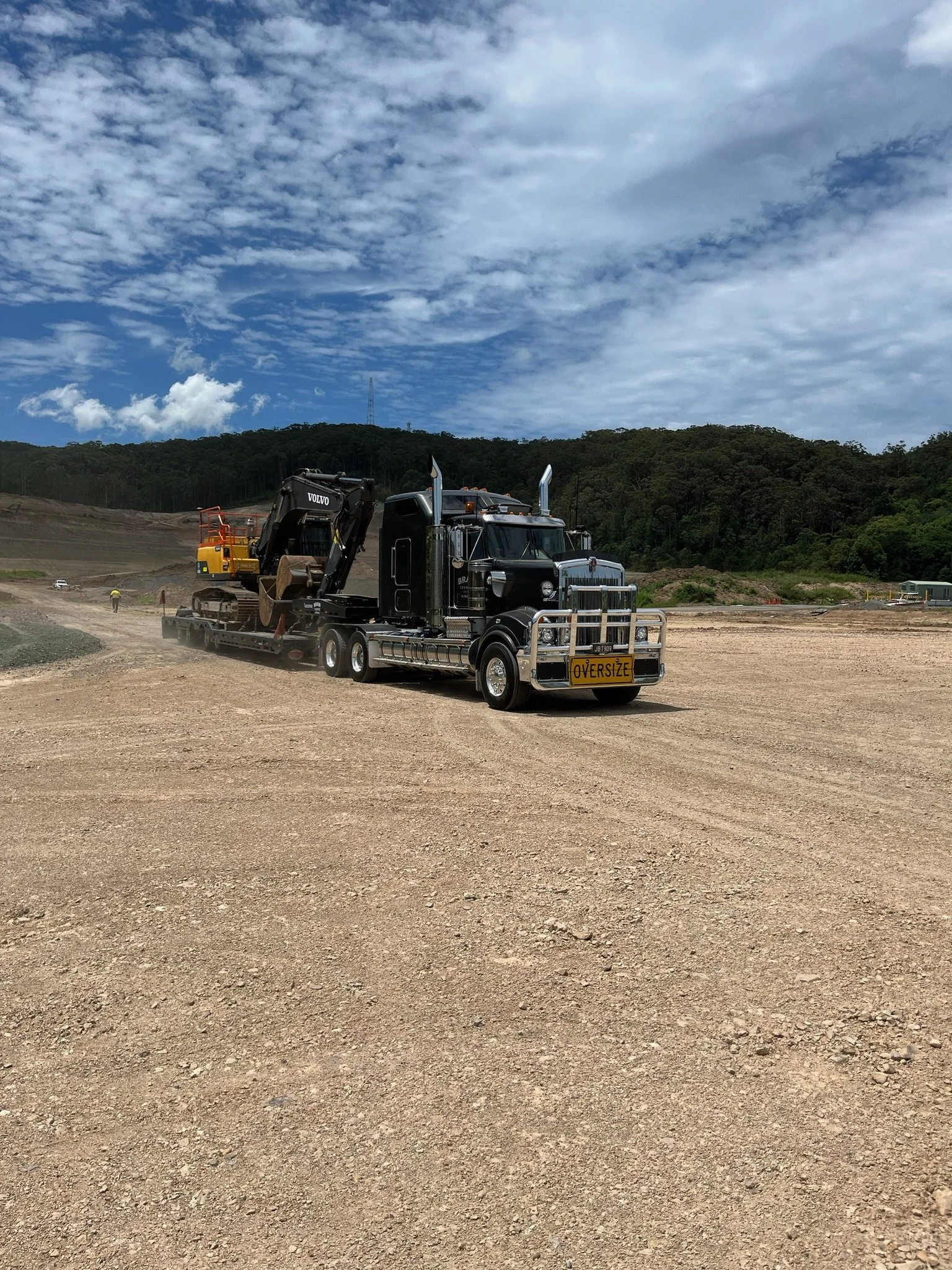 A black semi-truck with an oversized load sign on the front, carrying a yellow excavator on a flatbed trailer, on a dirt construction site with hills and trees under a partly cloudy sky.