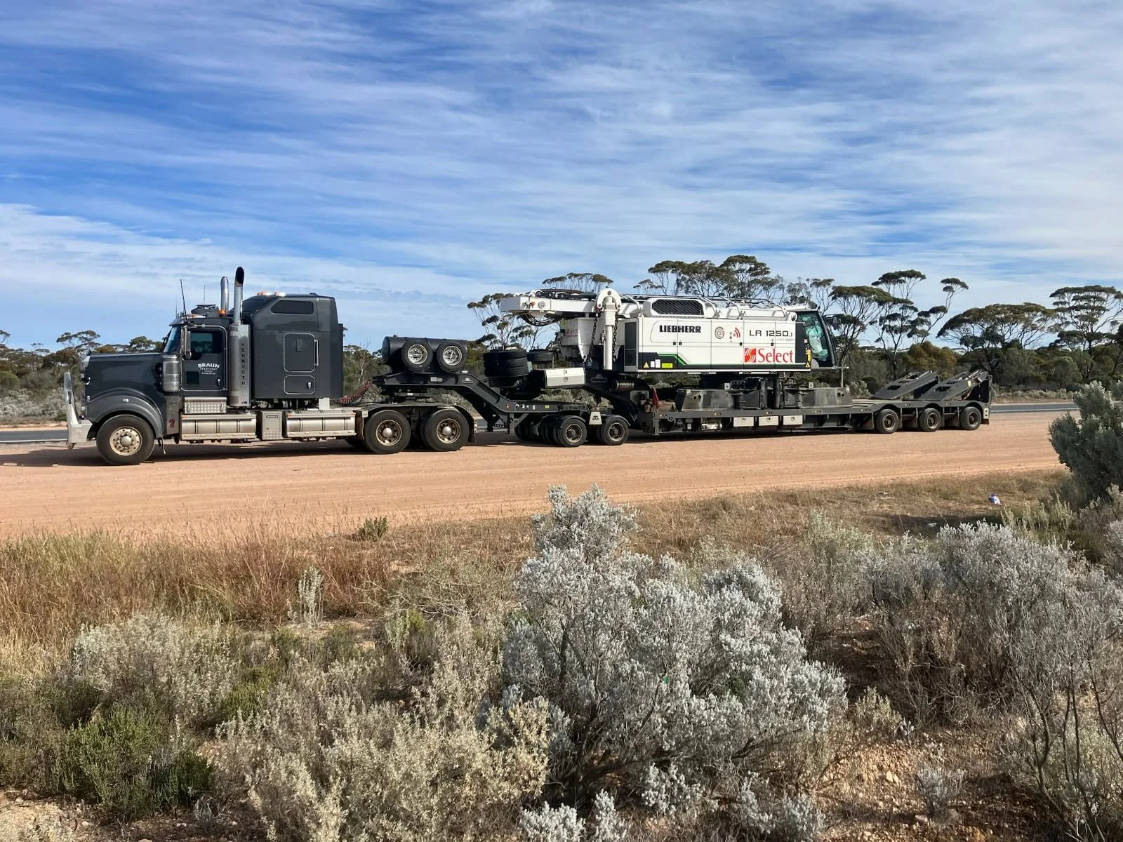 A large semi-truck is transporting construction equipment across a wide dirt road in a semi-arid landscape with sparse trees and bushes under a partially cloudy sky.