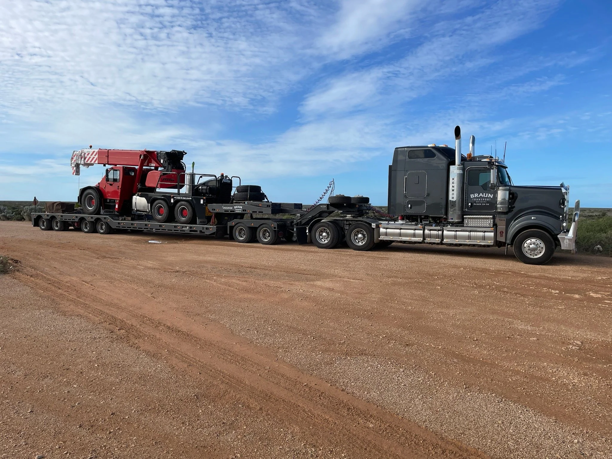 A large black semi-truck parked on a dirt road, carrying a flatbed trailer with a red motor grader and other equipment, against a blue sky with scattered clouds.