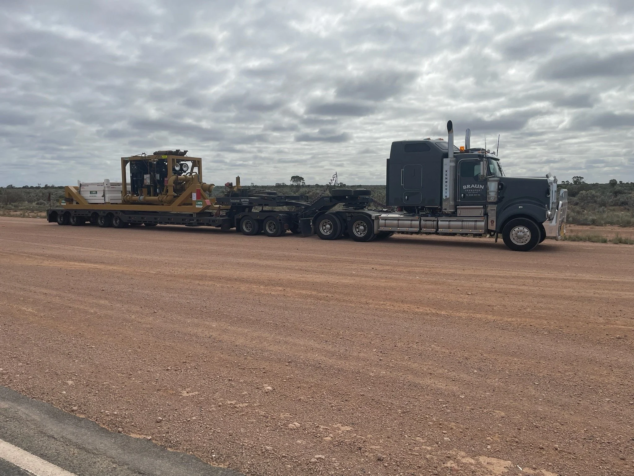 A semi-truck carrying heavy machinery on a flatbed trailer on a dirt road under a cloudy sky.