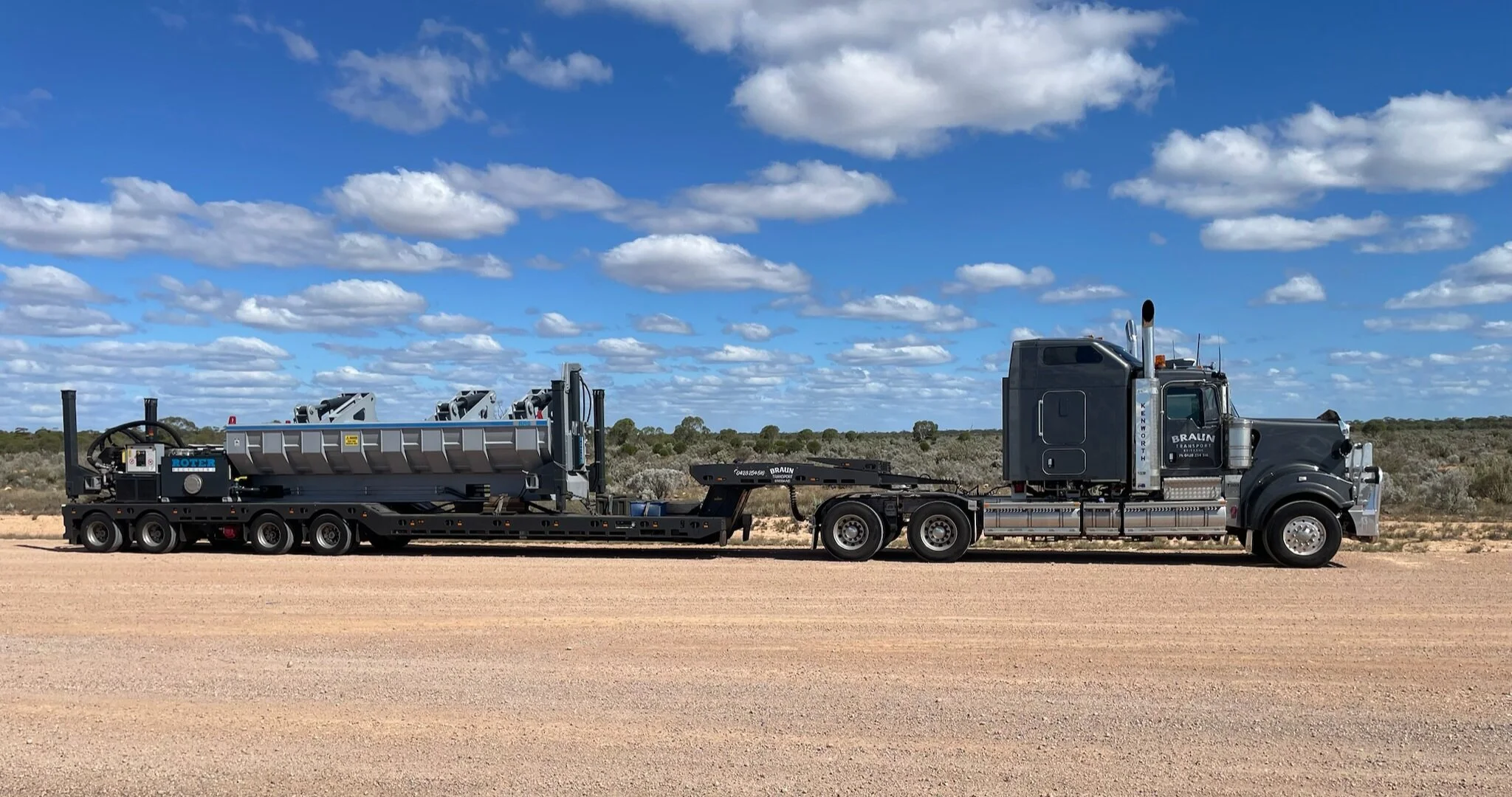 A large black semi-truck parked on a dirt road in a flat, open landscape under a partly cloudy sky. The truck is hauling industrial equipment on a flatbed trailer.